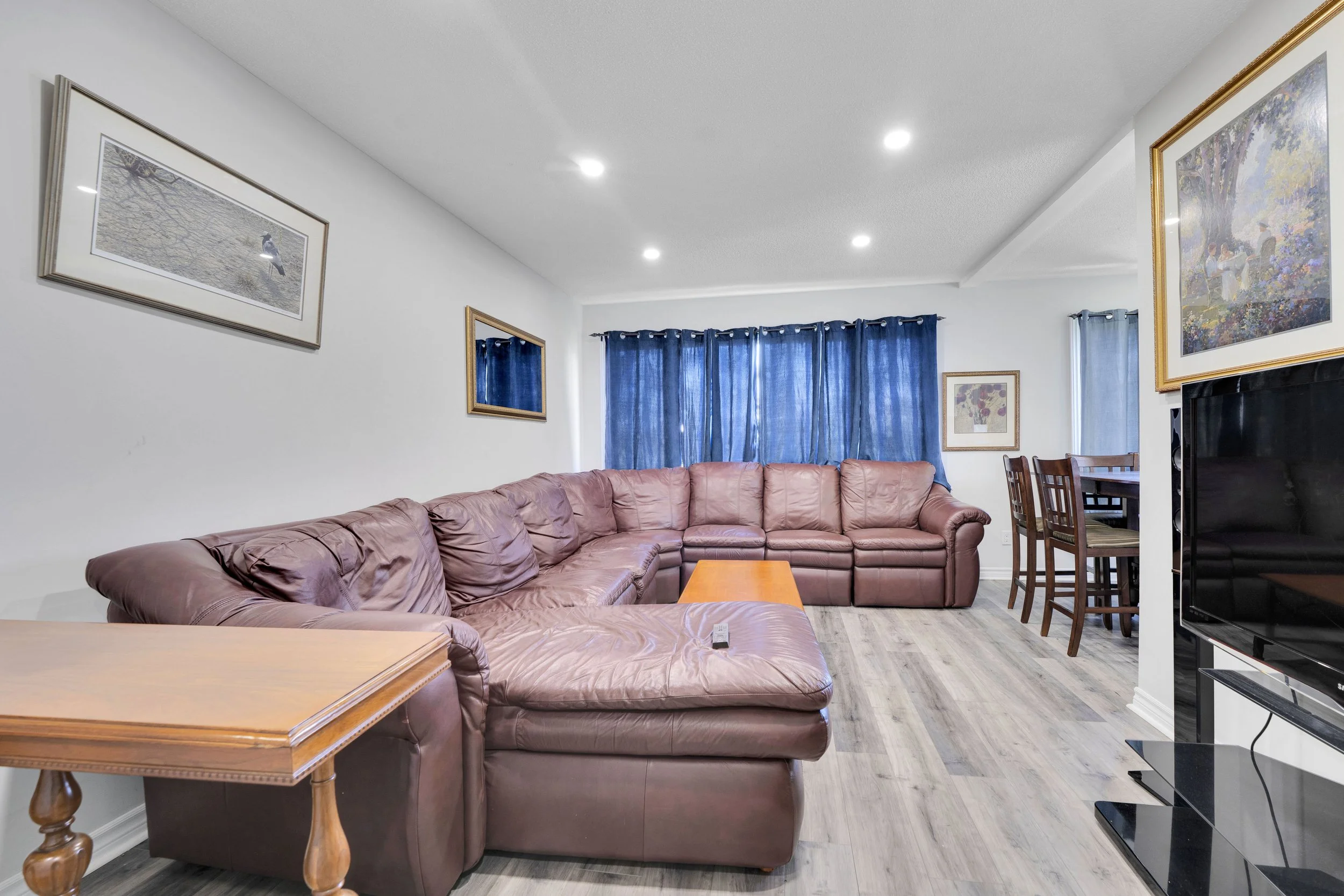 Ottawa Real Estate Media Services  Living room with brown leather sectional sofa, wooden side table, television, and blue curtains on windows, with framed artwork on white walls.