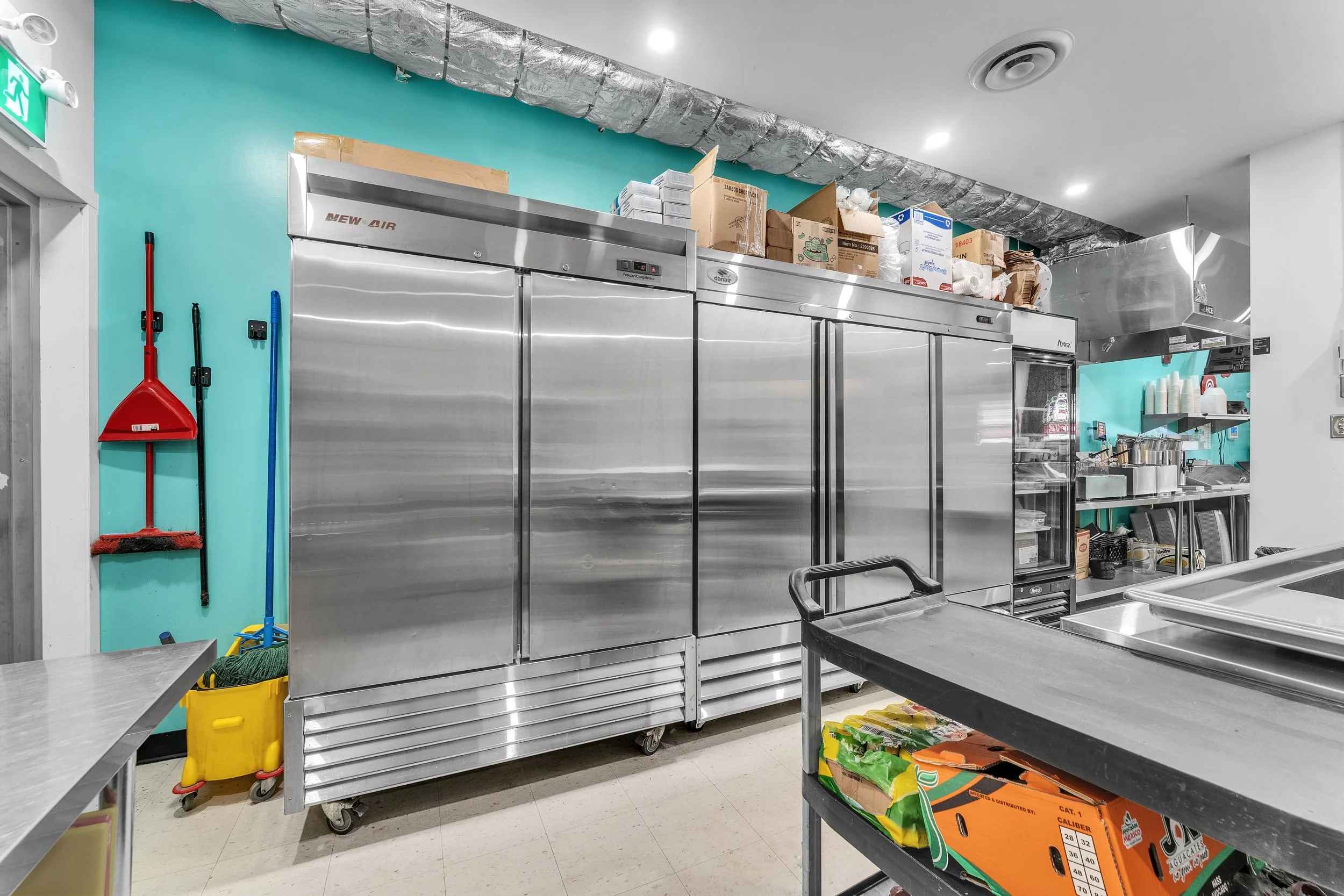 Commercial kitchen storage area with large stainless steel fridge, cleaning equipment like broom and mop, and a cart with supplies.