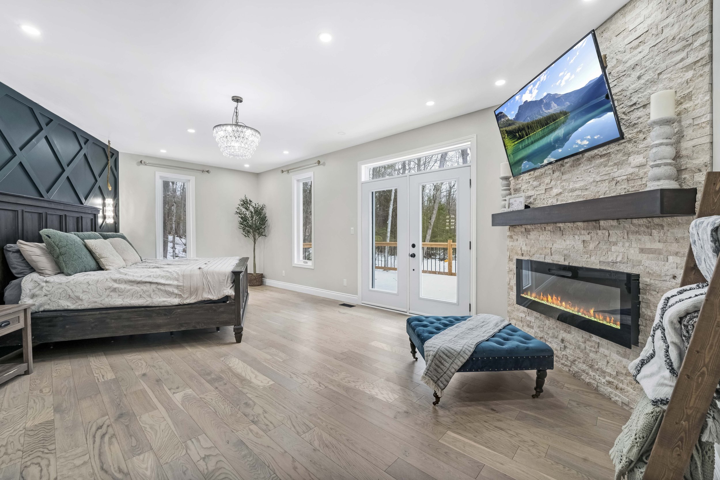NV3D Ottawa Real Estate Photography A bedroom with a black bed, green and white pillows, a fireplace with a TV mounted above it, a blue upholstered bench, a chandelier, and large windows with a view of a snowy outdoor area.
