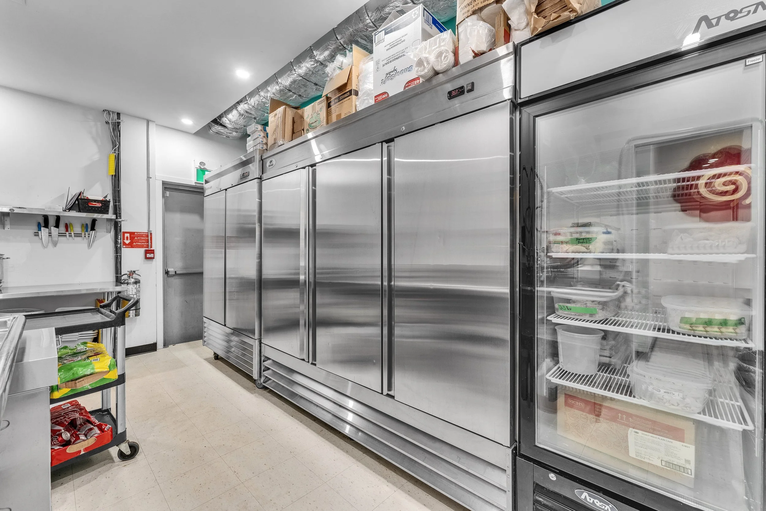Commercial walk-in refrigerator with stainless steel doors and glass door freezer showing various food items and slices of cake inside, with shelves and storage on top and on the nearby wall.