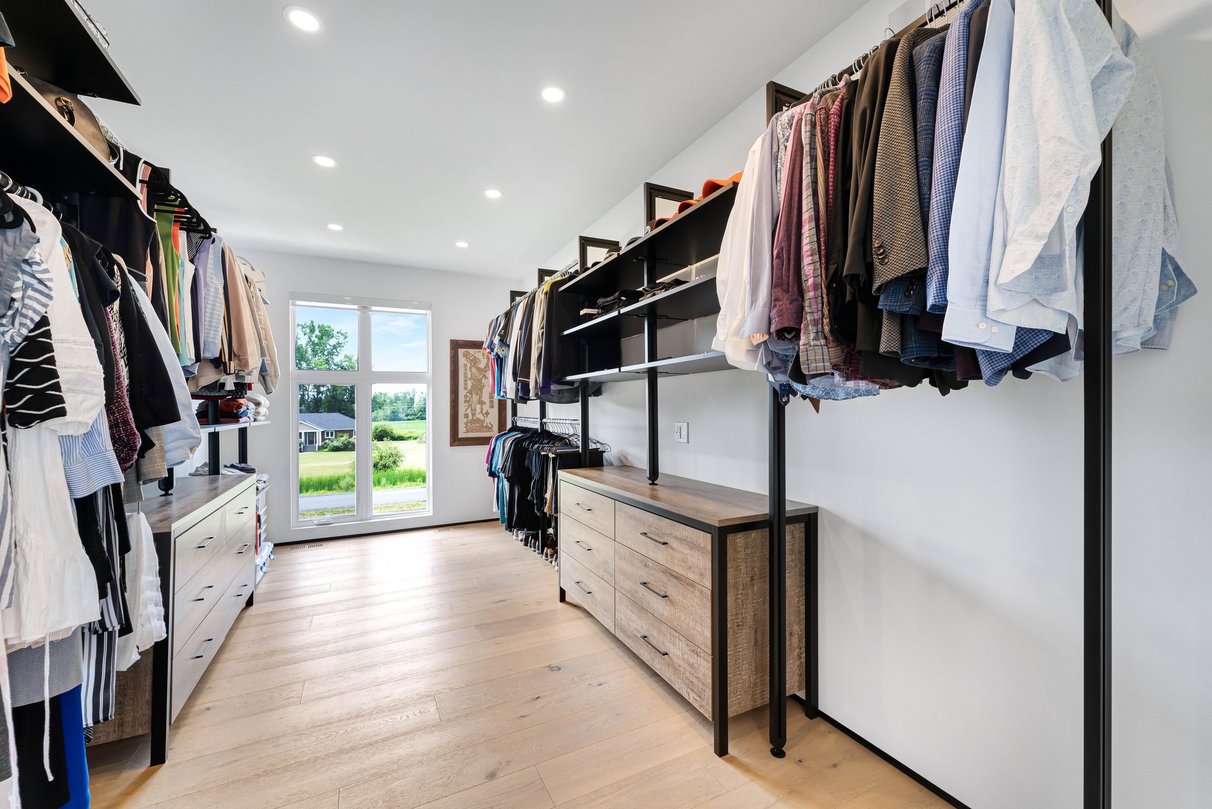 Modern walk-in closet with clothes on hangers and dressers, wooden flooring, and a window with a scenic view.