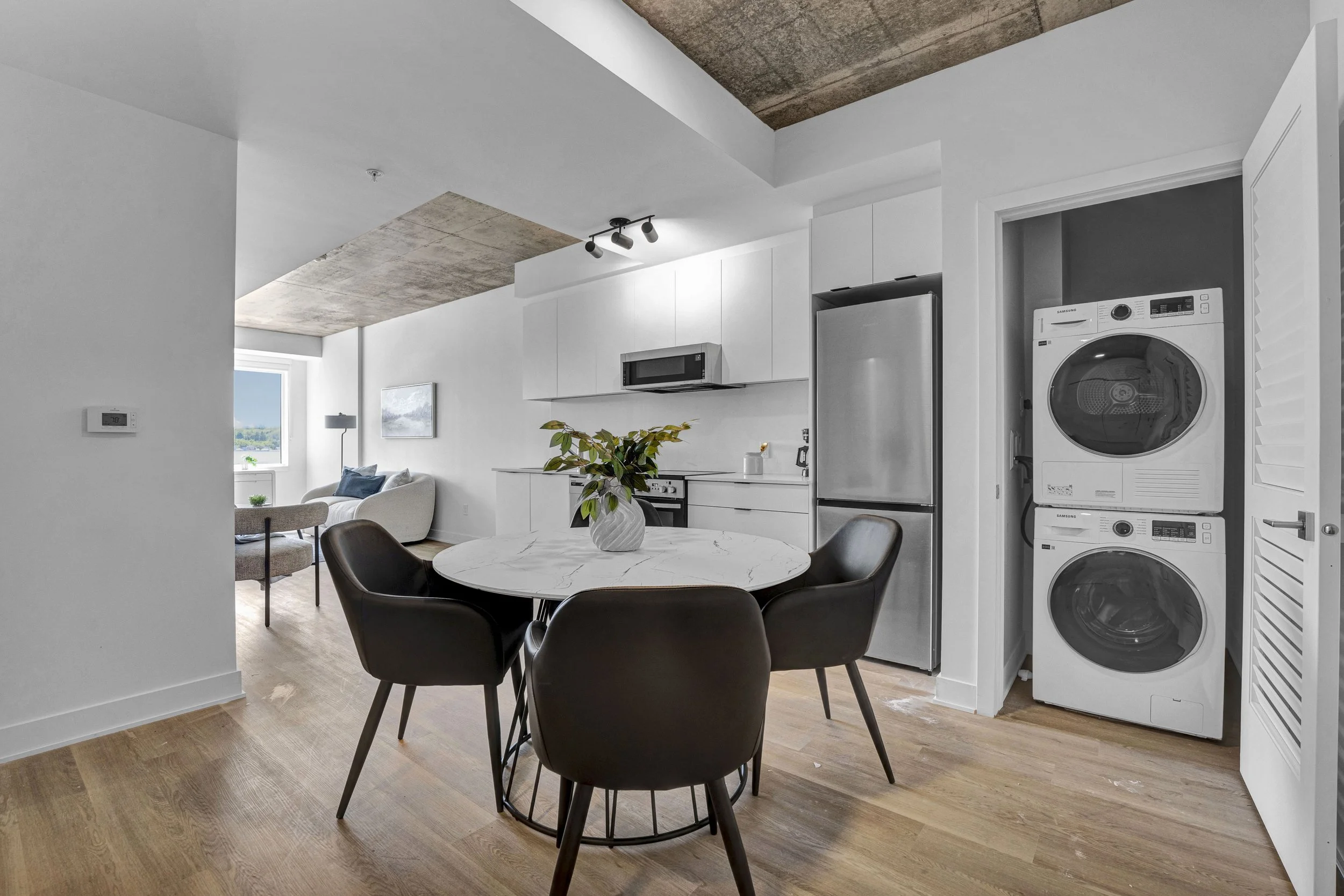 Open-concept living room and kitchen with white walls and ceiling, wooden flooring, and modern appliances including a stacked washer and dryer, a refrigerator, and a microwave. A round dining table with black chairs and a plant in a white vase are in