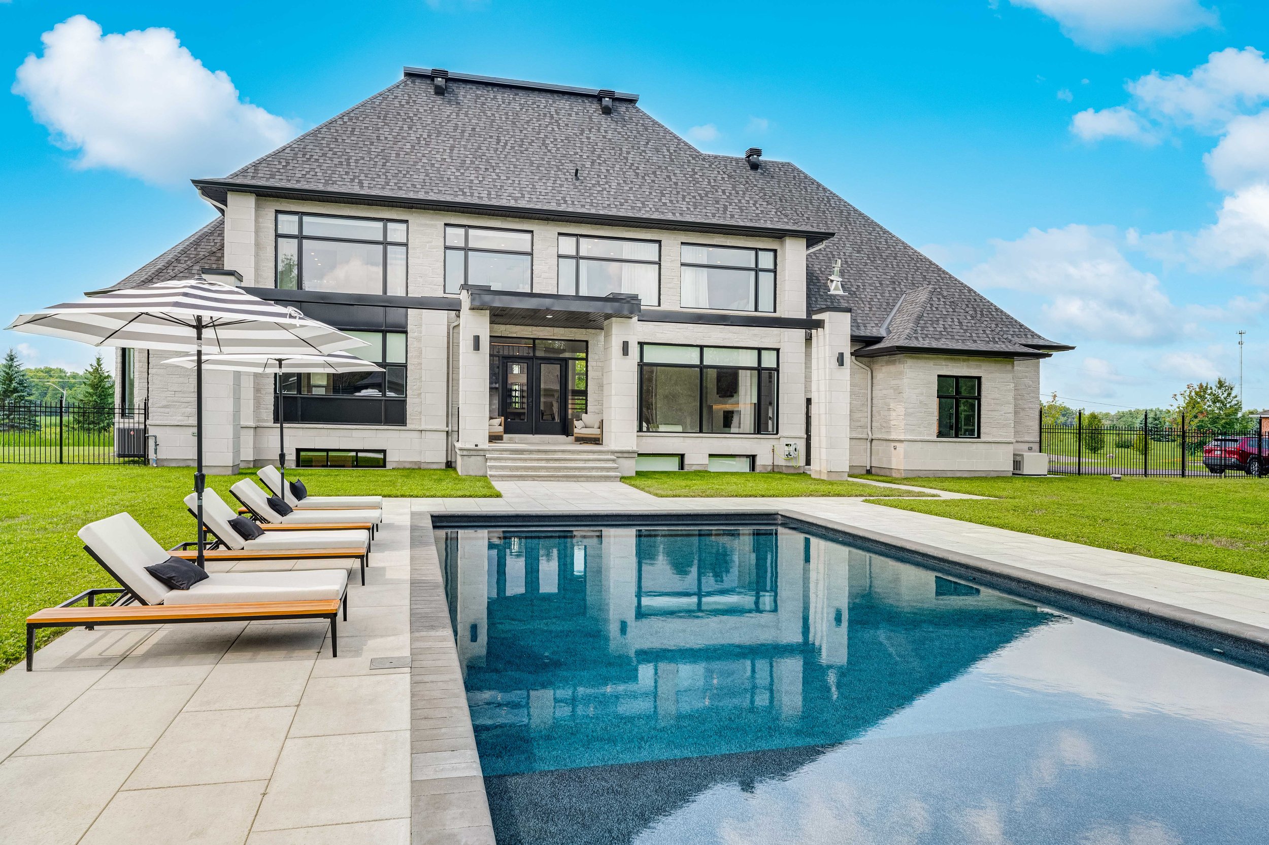 Luxury house with a pool, surrounded by lounge chairs and umbrellas, featuring large windows and a manicured lawn.