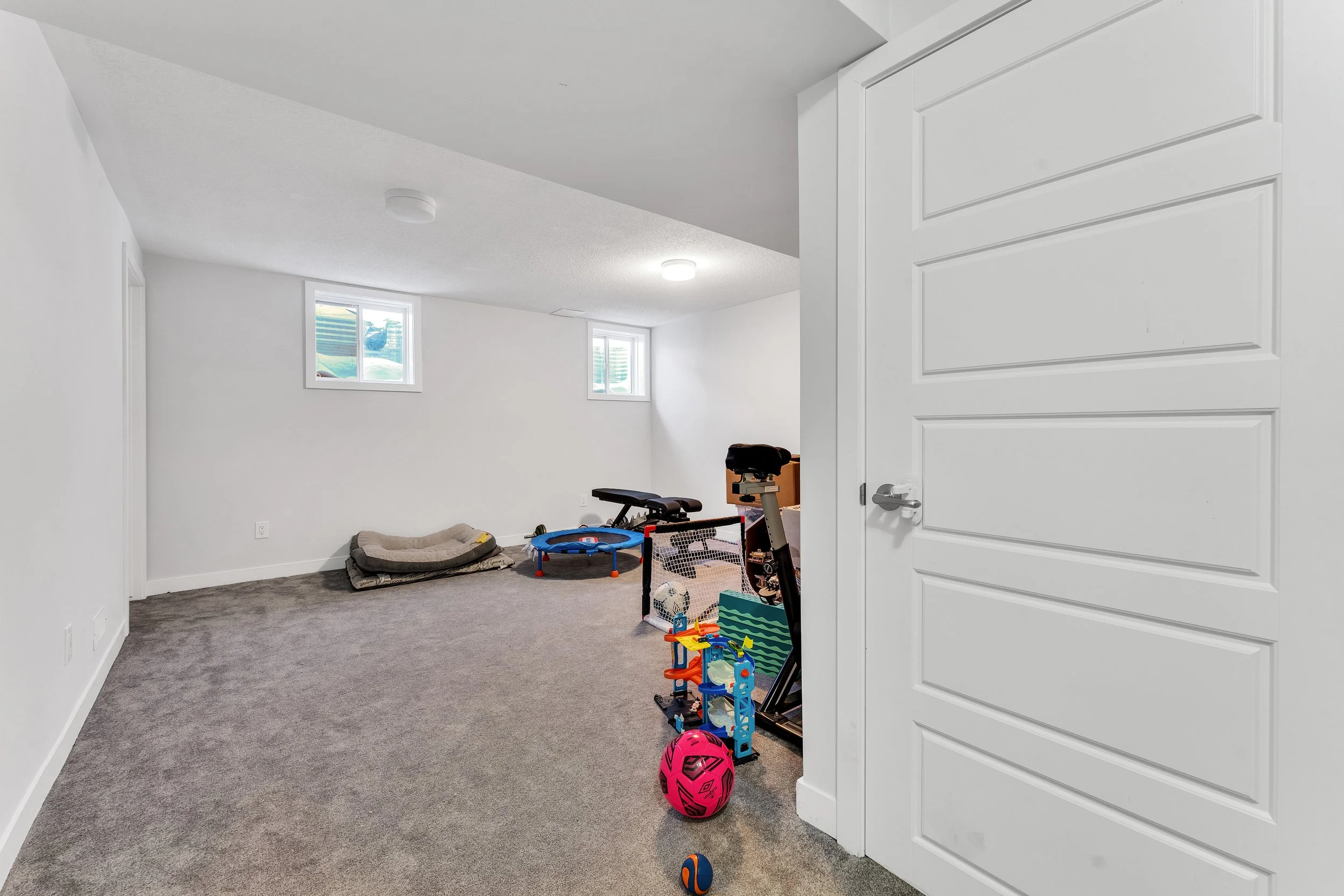 A basement room with white walls, two small windows, grey carpet, a pet bed, children's toys, a basketball, and exercise equipment.