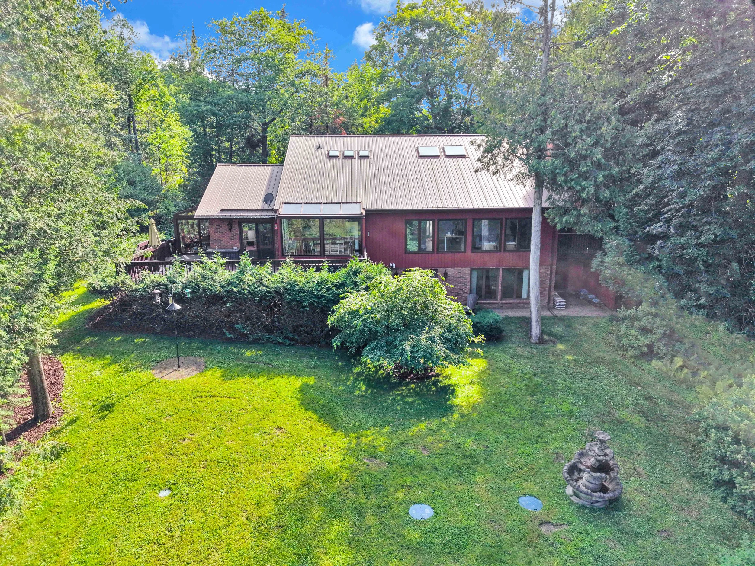 NVISION 3D Real Estate Media  A two-story house with a metal roof, large front windows, and a sunroom, surrounded by green trees and a manicured lawn under a blue sky with some clouds.