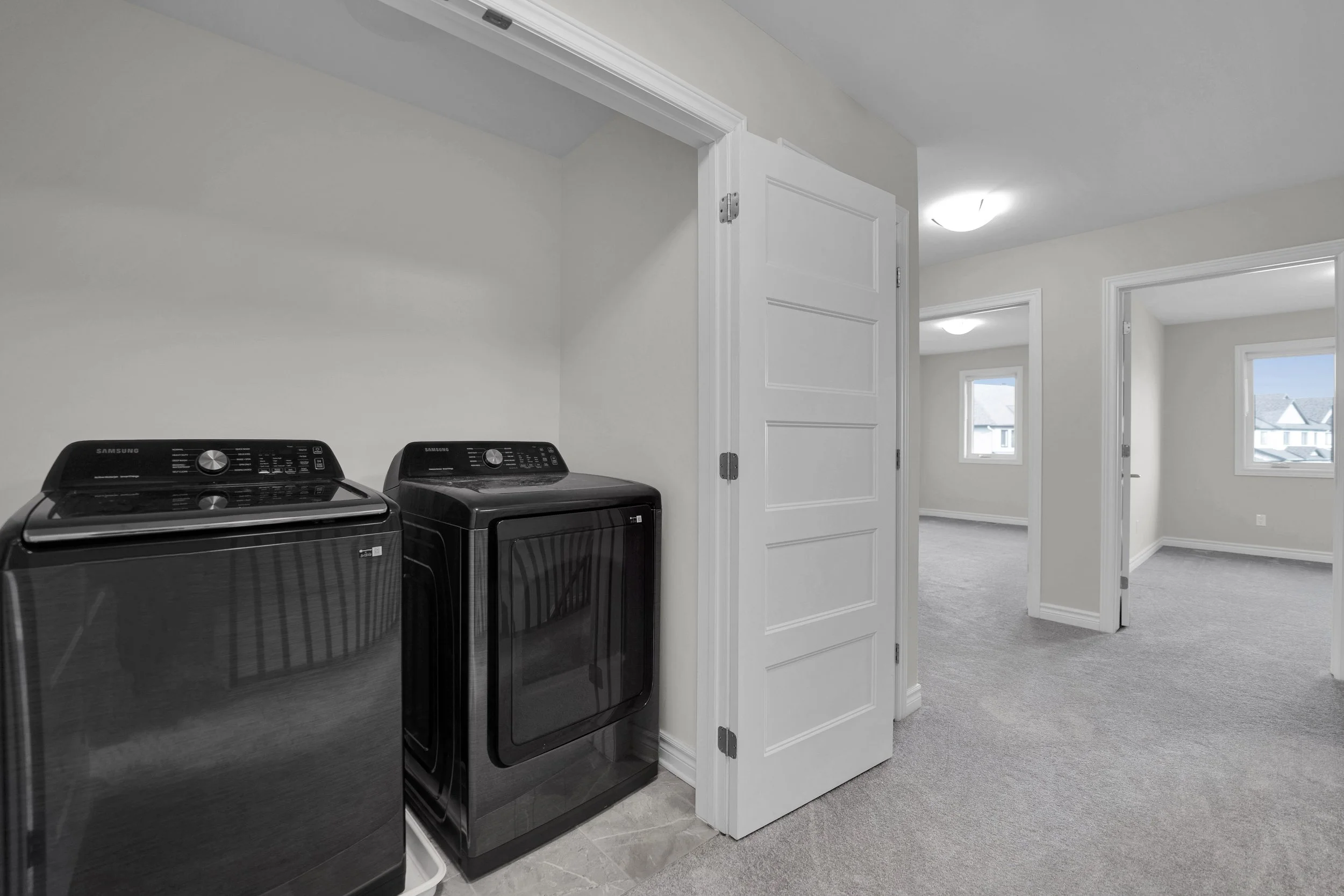 NVISION3D Real Estate Photography Ottawa  A laundry room with two black Samsung washer and dryer units against a white wall, open white door leading to a carpeted room with multiple windows showing a neighborhood view.