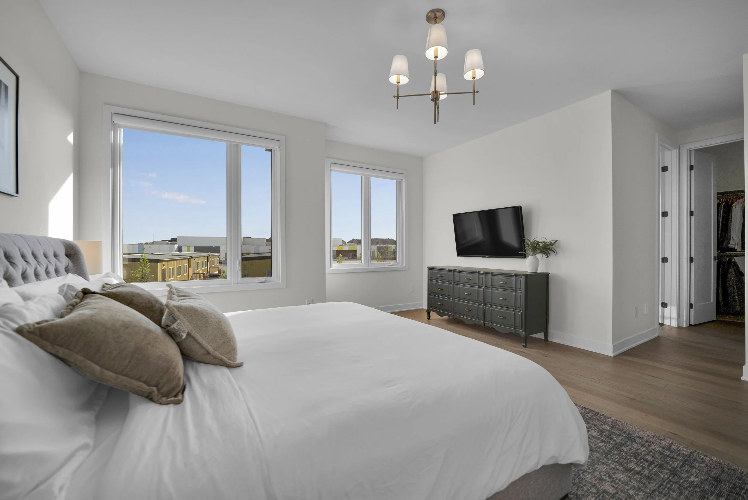 A bedroom with a large bed with beige pillows, a black dresser with a white vase and green plant, a flat-screen TV, and multiple windows letting in natural light, with a view of modern buildings outside.