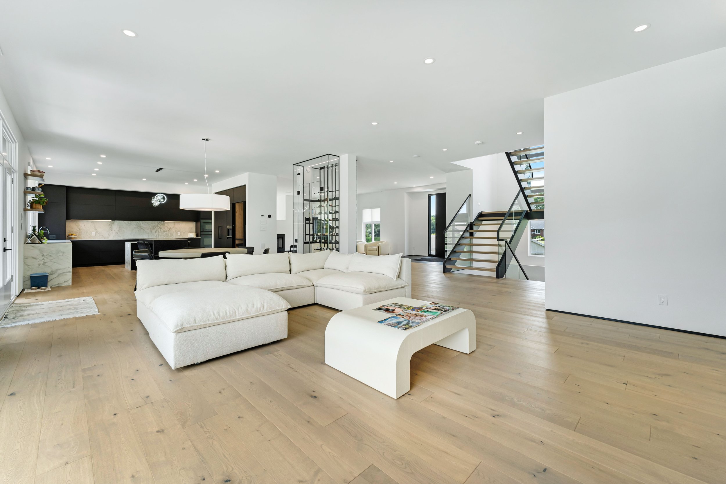 Spacious modern living room with beige sectional sofa, wooden floors, open kitchen, large windows, minimalist décor, and a staircase.