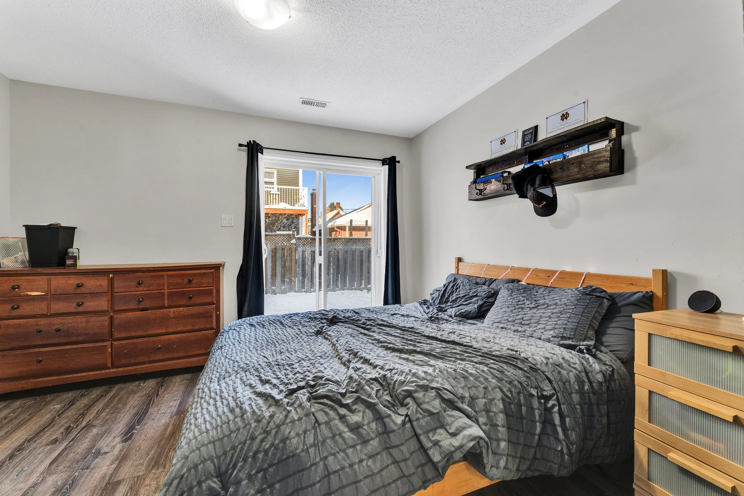 Bedroom with a bed, wooden dresser, sliding glass door with black curtains, and a wooden shelf on the wall holding picture frames and a cap.