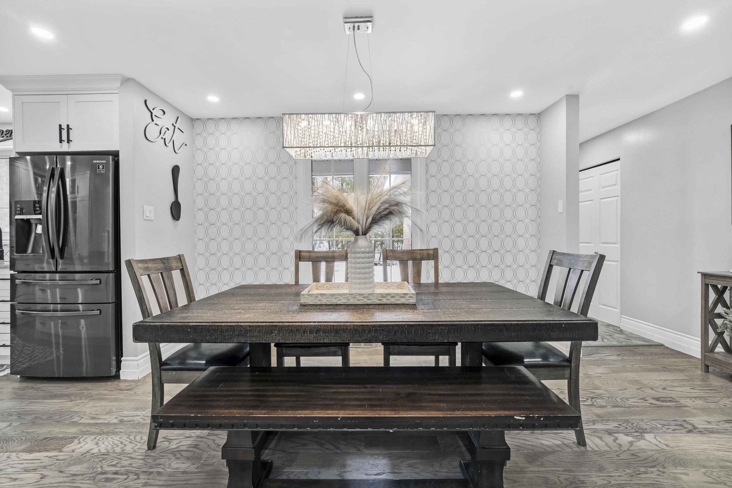 Real Estate A dining room with a dark wood table, six matching chairs, and a bench. A decorative tray with a vase and pampas grass centerpiece is on the table. Behind, a window with a geometric-patterned wallpaper and hanging light fixture above.