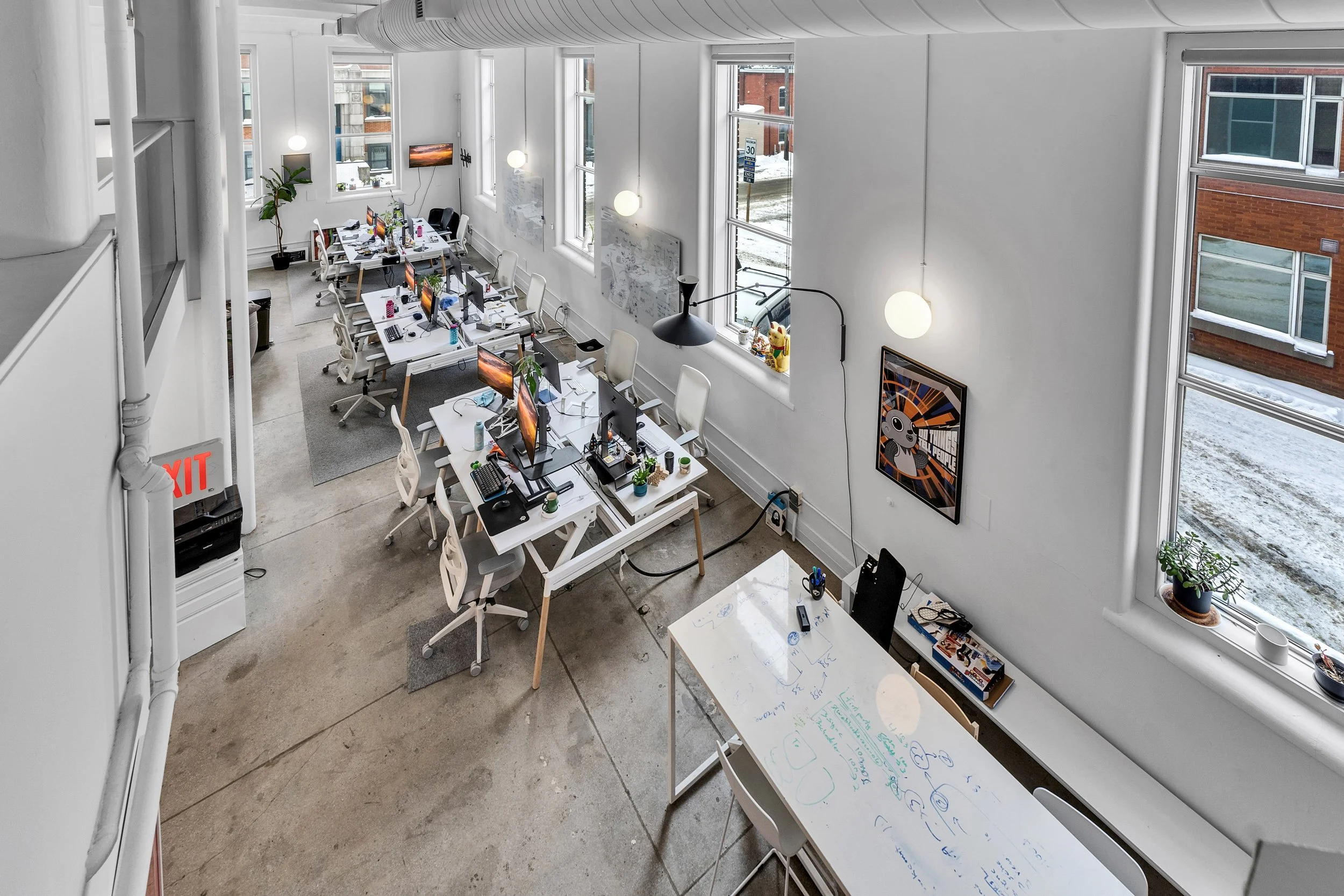 An open-plan office with multiple workstations, computers, and office supplies. Large windows with snow outside, minimalistic decor, white walls, hanging lights, and an abstract framed artwork.