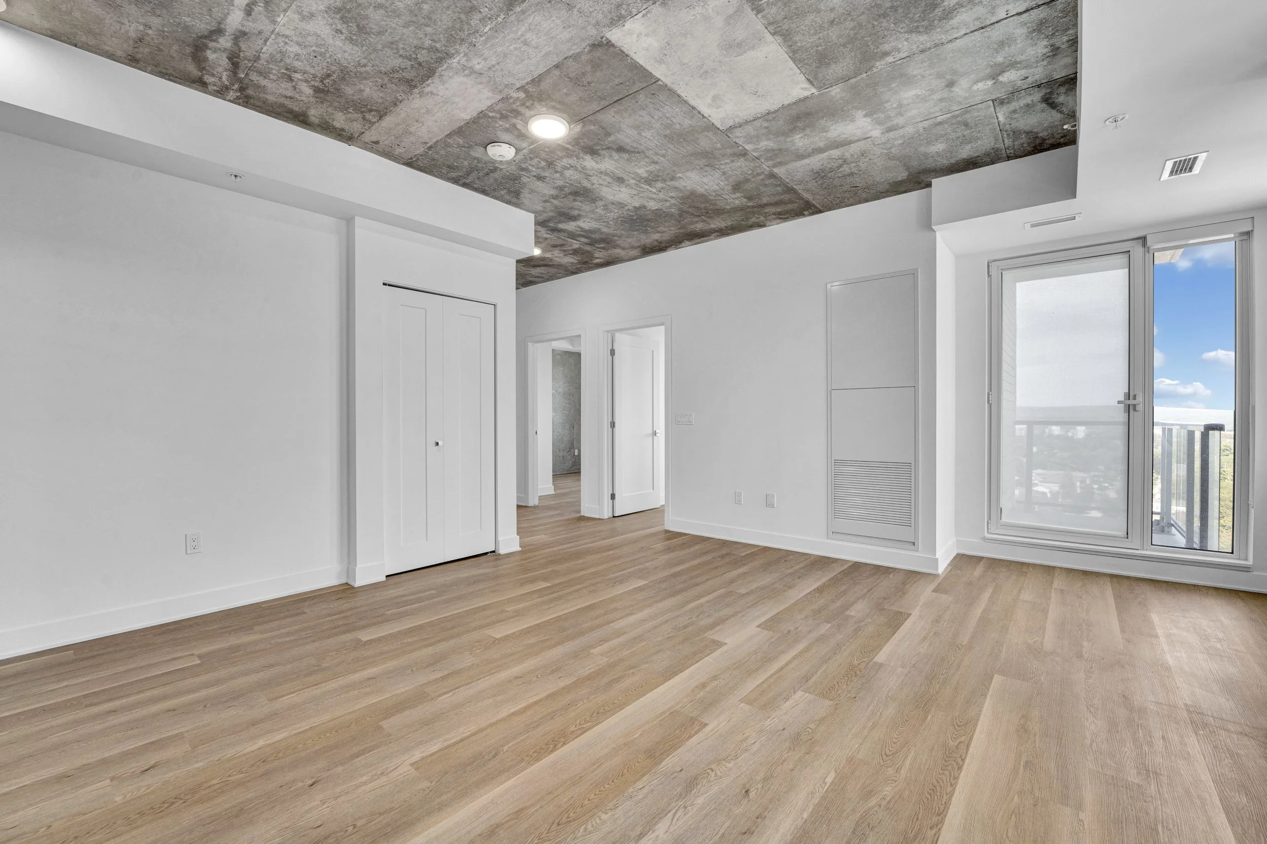 Empty living space with white walls, large window with a city view, wood flooring, and a textured concrete ceiling.