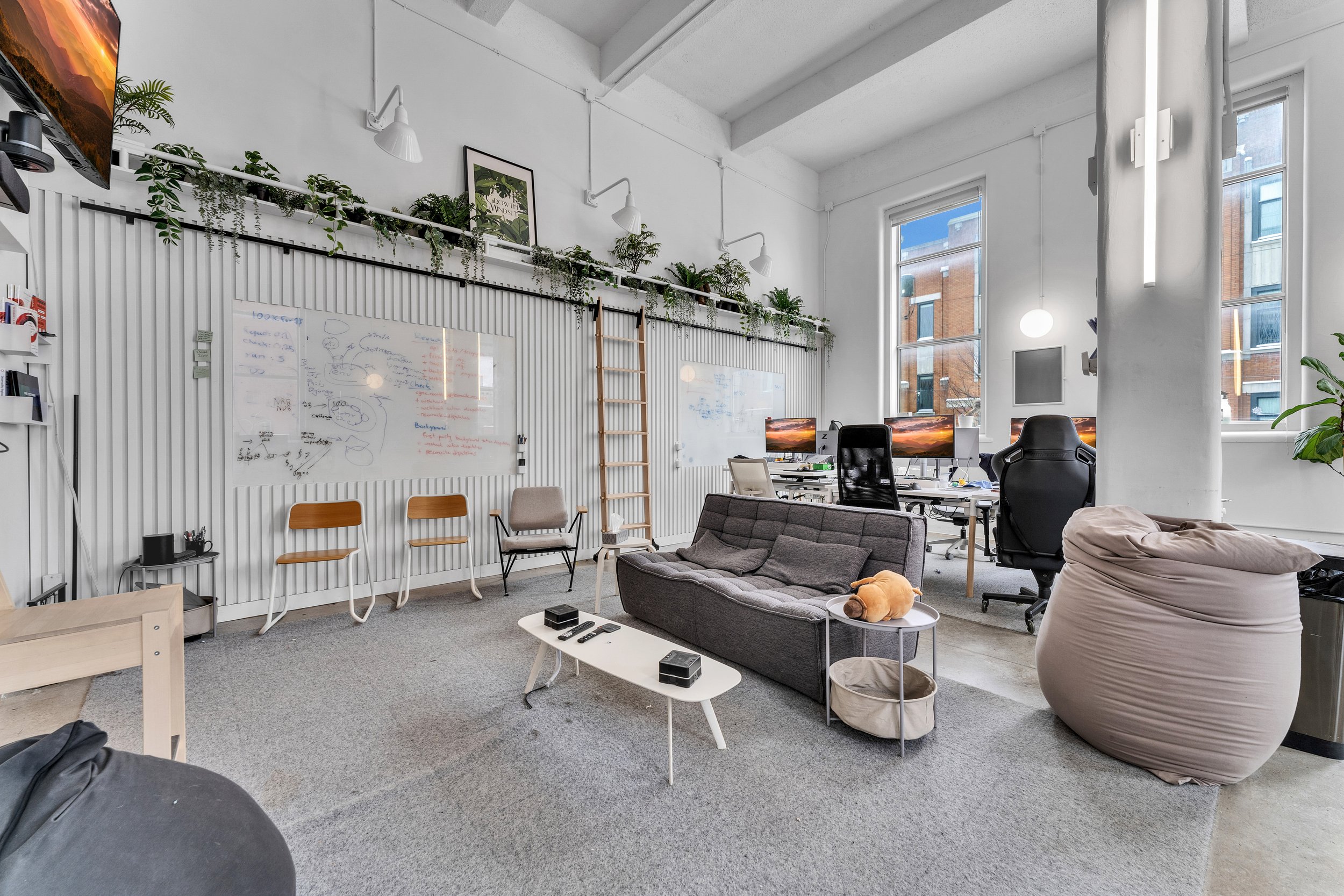 A modern office with white walls, large windows, a gray carpet, and multiple desks with computer monitors. There is a gray sofa with a pillow and a plush toy, a beige bean bag chair, and several chairs along the wall with whiteboards. Potted plants a