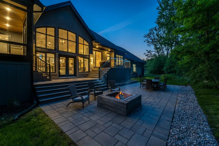 A modern house with large windows and a lit patio area, featuring a fire pit, wicker chairs, and an outdoor dining table, surrounded by trees under an evening sky.