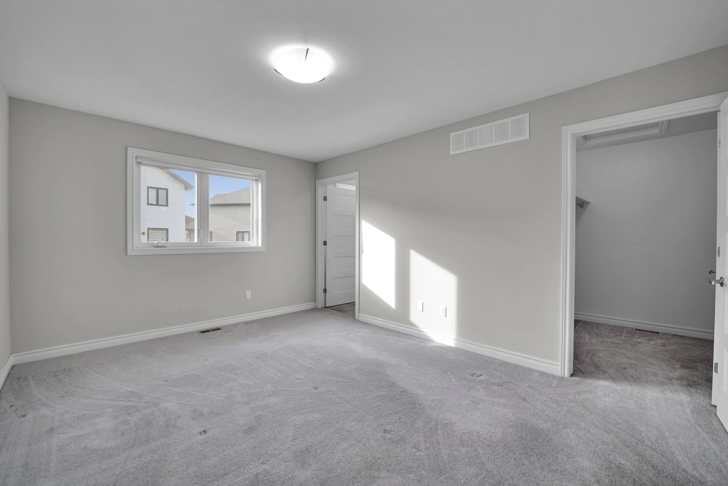 NVISION3D Real Estate Photography Ottawa  Empty room with beige carpet, white walls, and a window showing neighboring houses. There is a ceiling light fixture, a door leading to another room, and a closet with an open door.