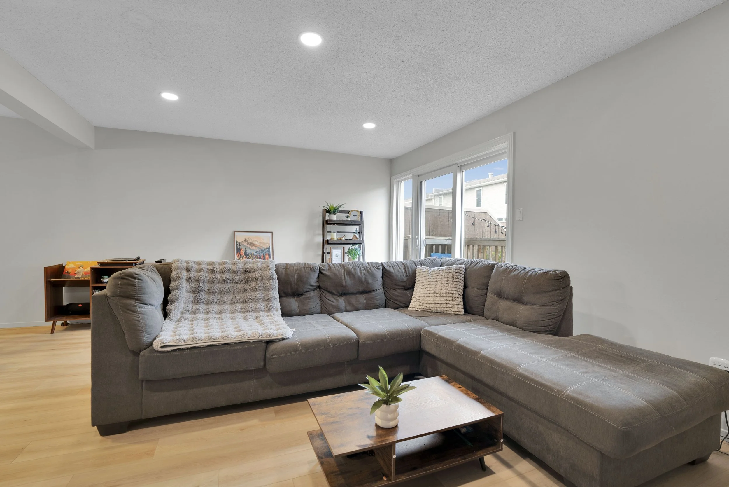 Ottawa Real Estate Video Services  Living room with gray L-shaped sectional sofa, wooden coffee table with small green plant, art print, window with view of neighboring houses and fence, bookshelf with decorative items, and hardwood flooring.