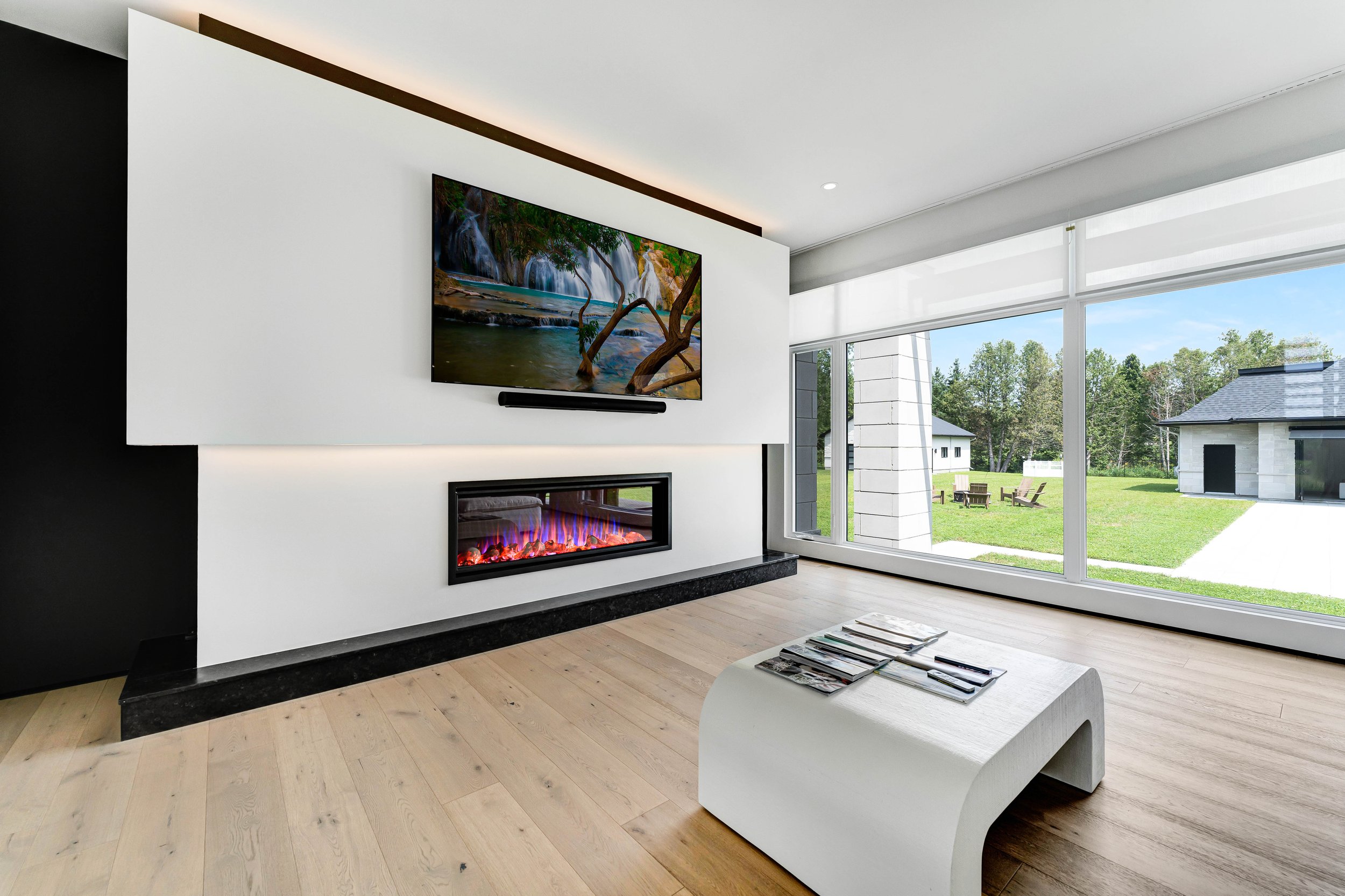Modern living room with a wall-mounted TV above a sleek electric fireplace, a minimalist white coffee table, wood flooring, and large windows overlooking a green backyard.