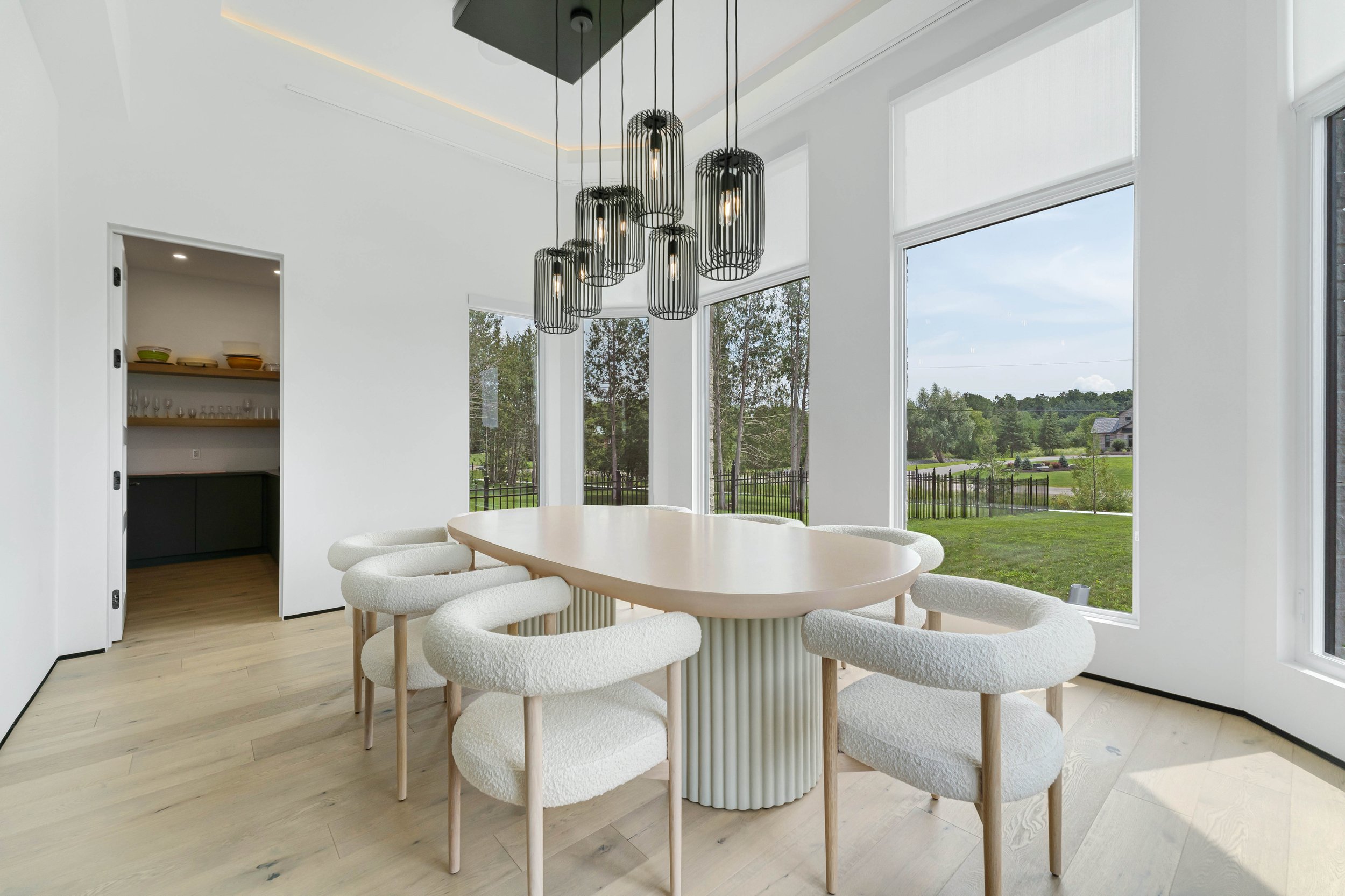 Modern dining room with a sleek light wood table, surrounded by six decorative chairs, and pendant lights hanging above. Large windows reveal a scenic outdoor view.