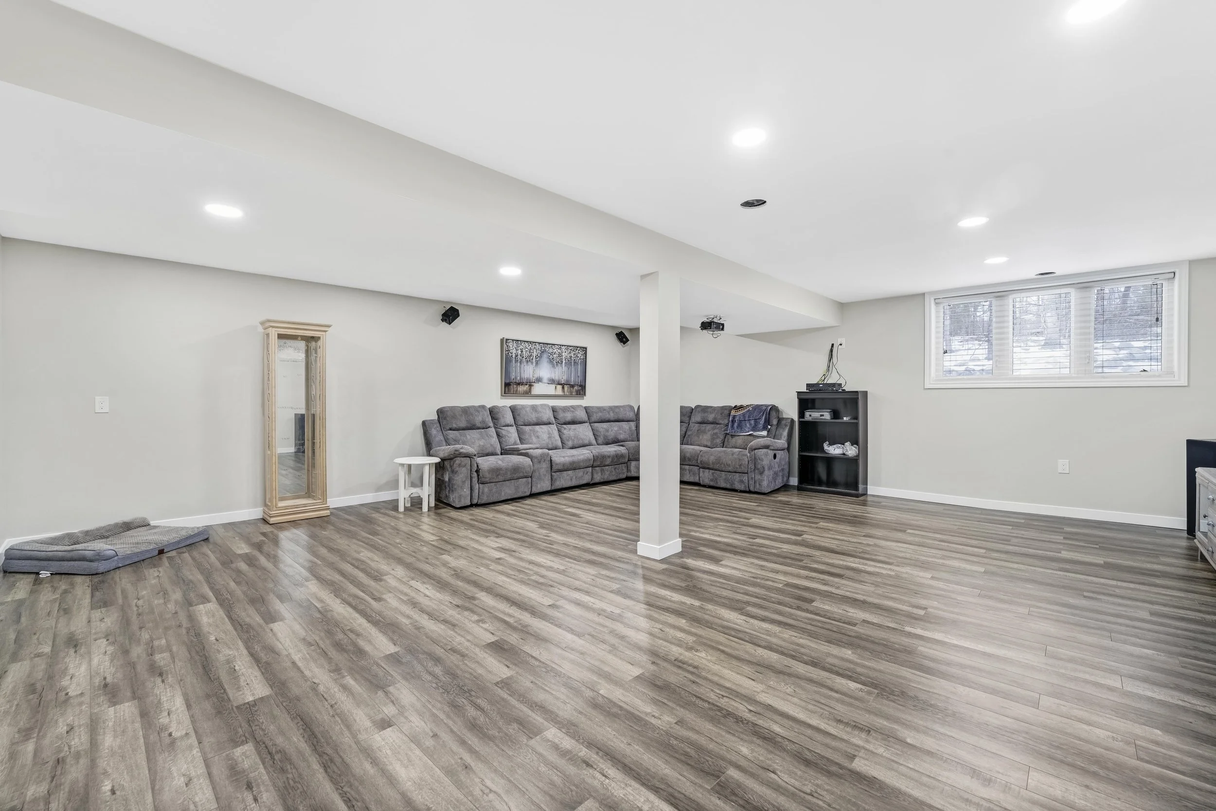 Spacious basement living area with gray sofas, a corner TV stand, a window with blinds, and wood-look flooring.