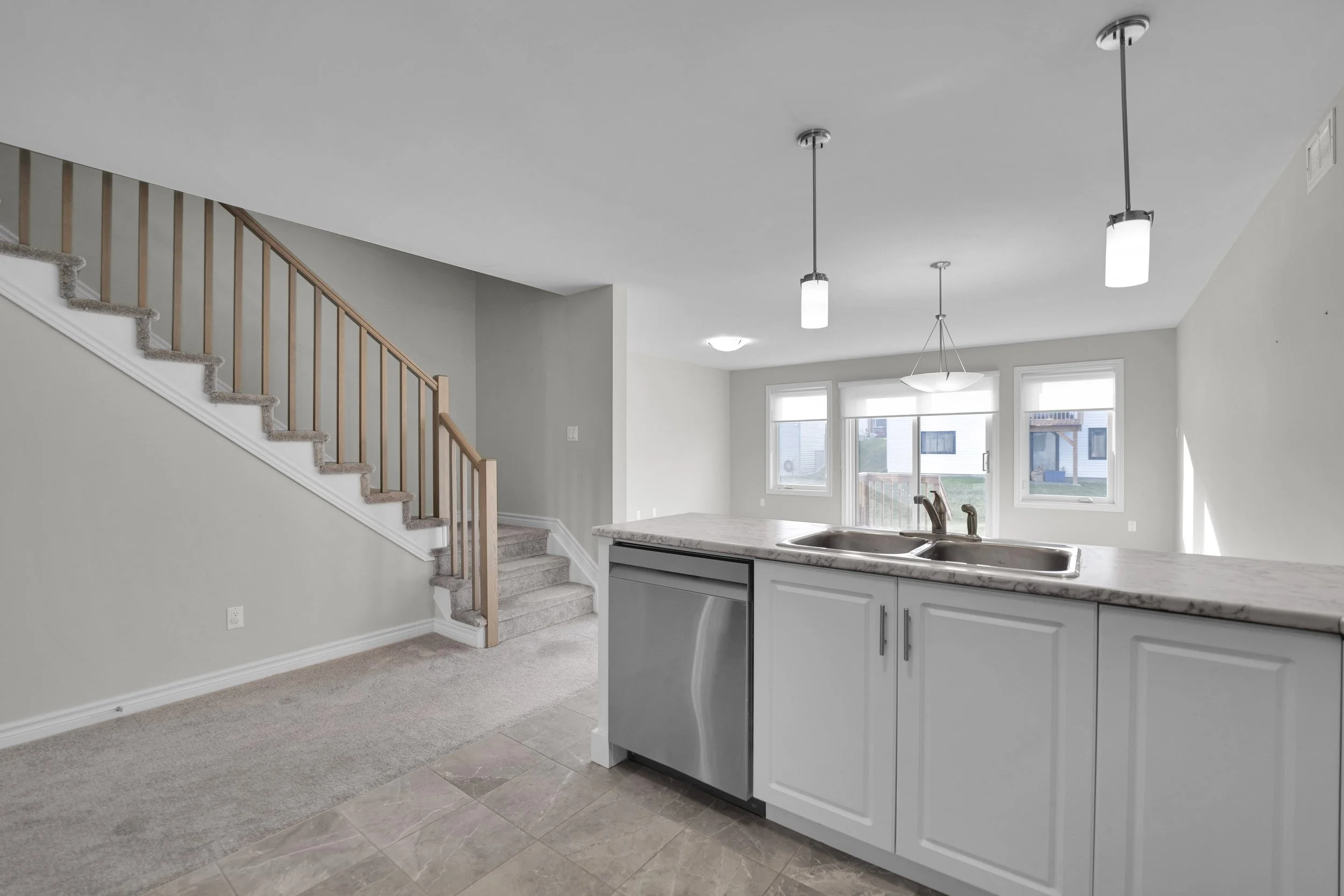 NVISION3D Real Estate Photography Ottawa  Interior of a modern home with a kitchen and staircase. The kitchen features a marble countertop, stainless steel sink, white cabinets, and pendant lighting. Large windows let in natural light.