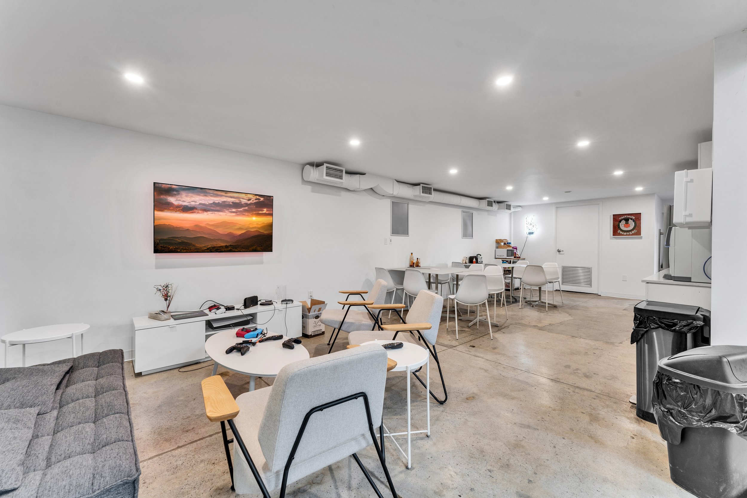 A spacious, modern communal area with white walls, seating, a wall-mounted TV showing a sunset over mountains, a small table with gaming controllers, and a dining area in the background with chairs and tables, some appliances, and a coffee station.