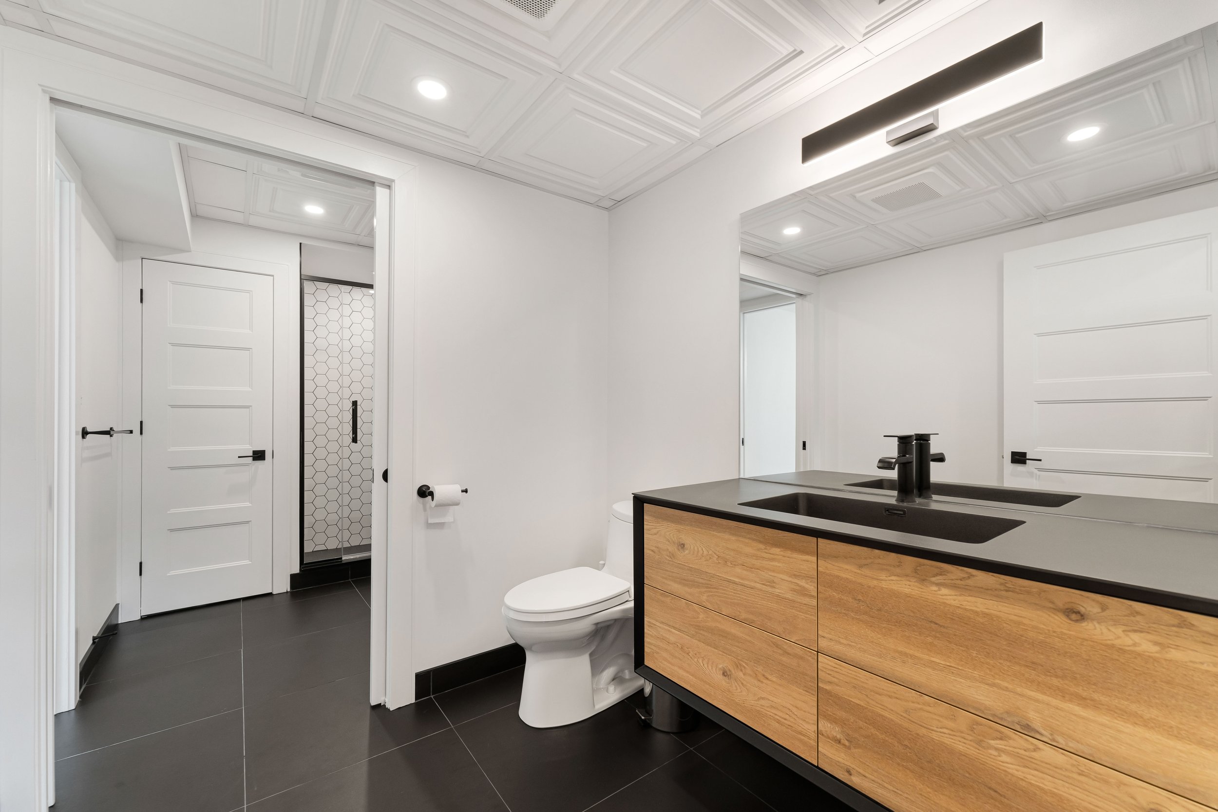 Modern bathroom with white walls, black tile floor, wooden vanity with black sink and faucet, large mirror, toilet, and shower with glass door and honeycomb tiles.