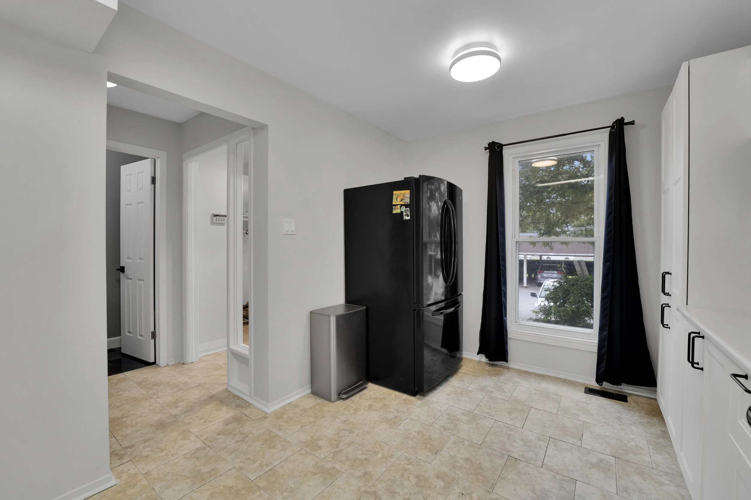 The image shows a section of a kitchen with beige tile flooring and white walls. There is a black refrigerator near the window, and black curtains hang on either side of the window. The window offers a view of trees and parked cars outside. There is 