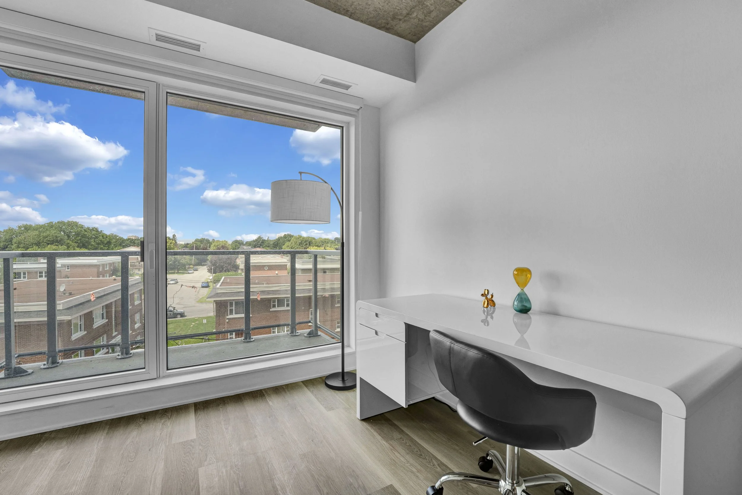 Modern interior room with white walls and large glass sliding doors leading to a balcony with a view of trees and buildings against a blue sky with clouds. There is a white desk with a black office chair, a tall white floor lamp with a drum shade, an