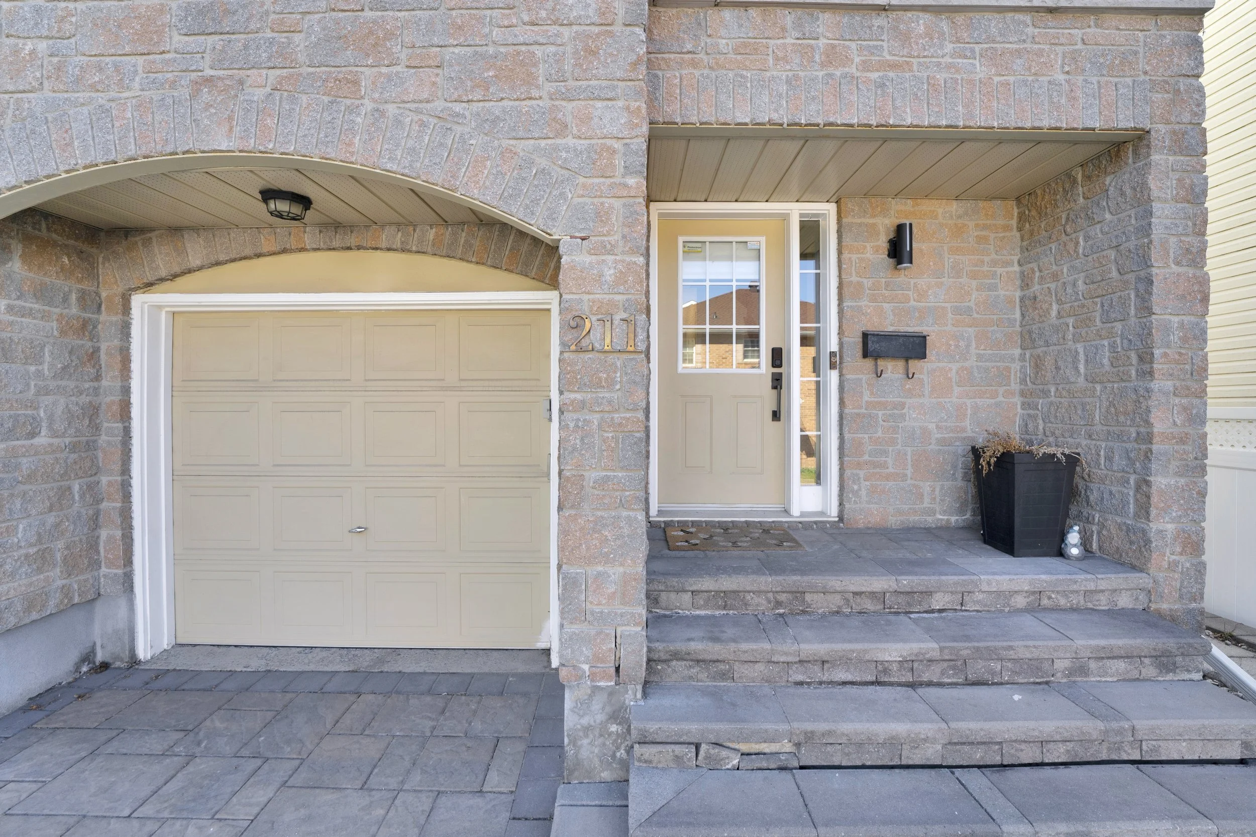 The image shows the front of a modern house with a brick facade, a small porch, and a garage. The house number 211 is displayed on the brick wall next to the front door. There is a black mailbox mounted on the wall next to the door, and a black outdo