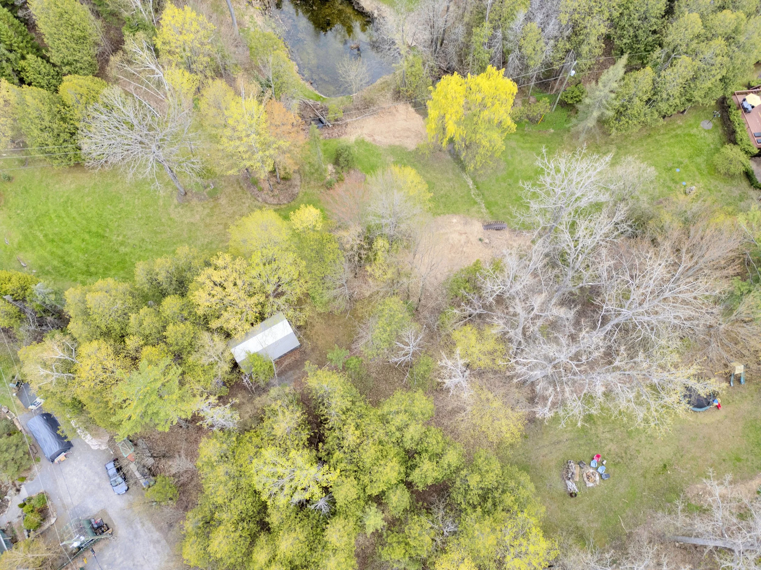 Ottawa Real Estate Media Services  An aerial view of a residential backyard with trees, grass, a small pond, some cars, and outdoor furniture.