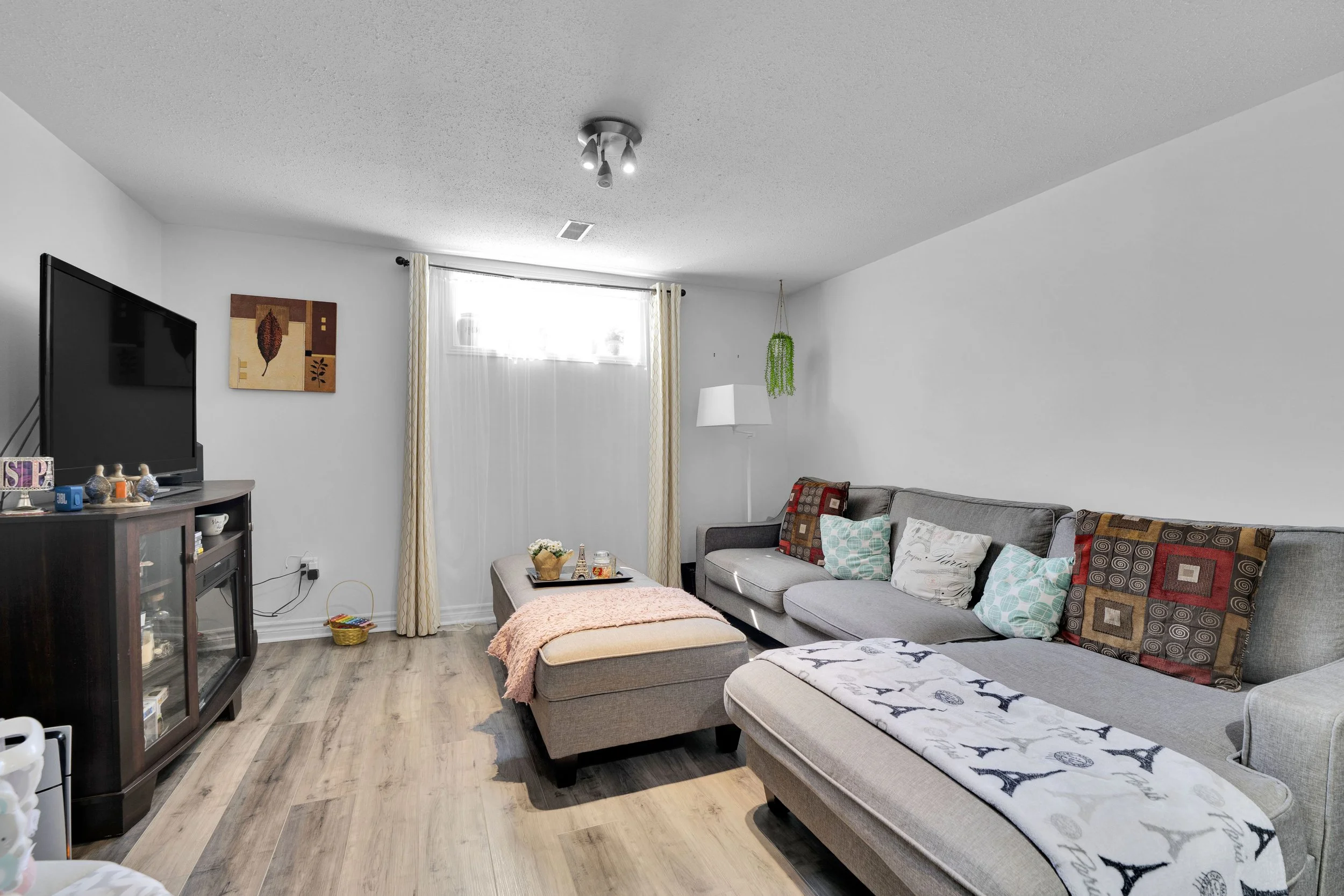 Ottawa Real Estate Media Services  Living room with gray sectional sofa, patterned throw pillows, wall art, window with curtains, floor lamp, and entertainment center with television.