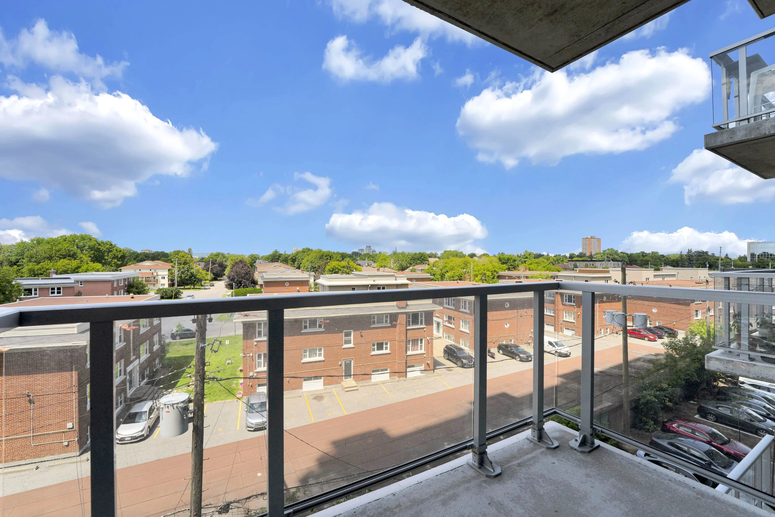 Ottawa Real Estate Photographer View from a balcony overlooking a parking lot surrounded by low-rise brick apartment buildings, trees, and a bright blue sky with scattered clouds.