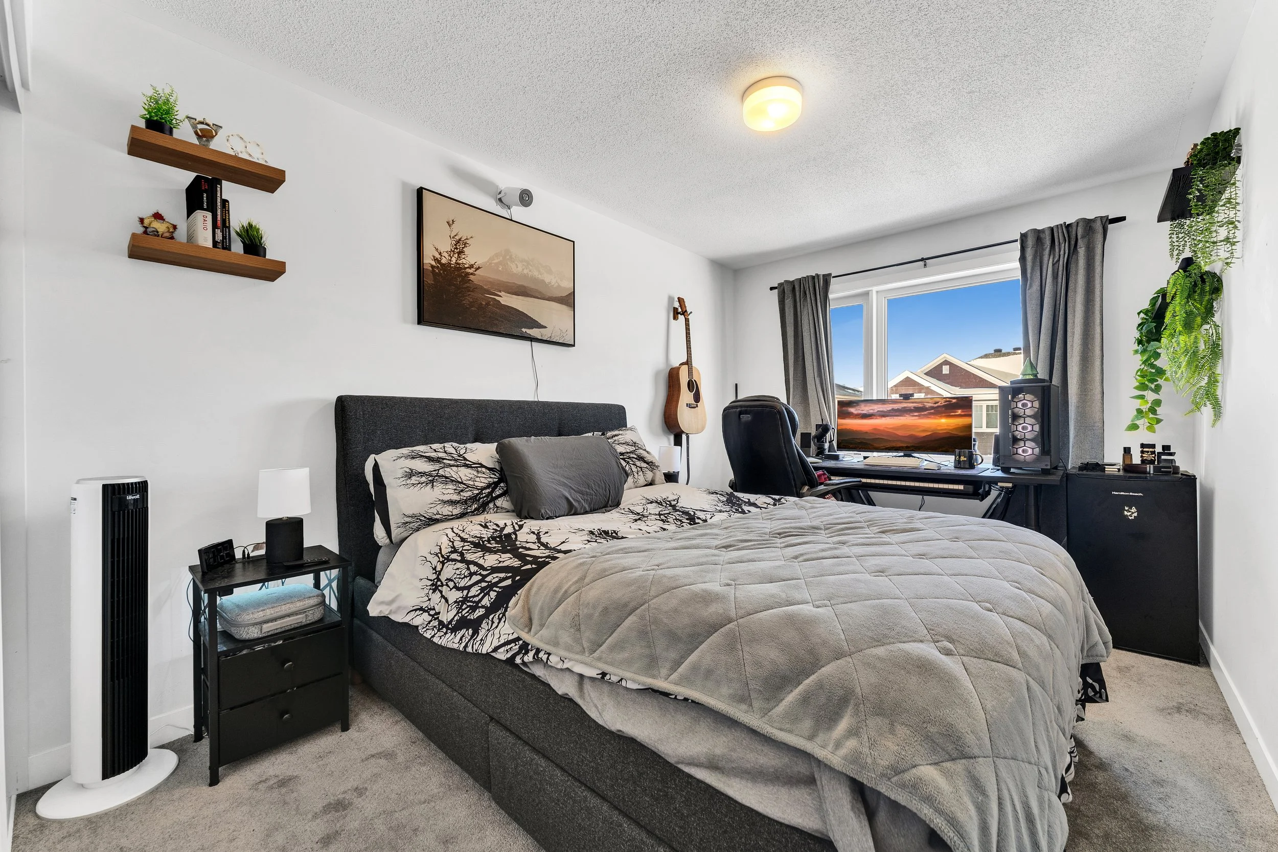 A bedroom with a bed featuring a tree branch design on the bedding, a nightstand with a lamp, a computer workspace with a monitor, keyboard, speakers, and a gaming tower, a guitar hanging on the wall, a window with curtains, hanging plants, and a sta
