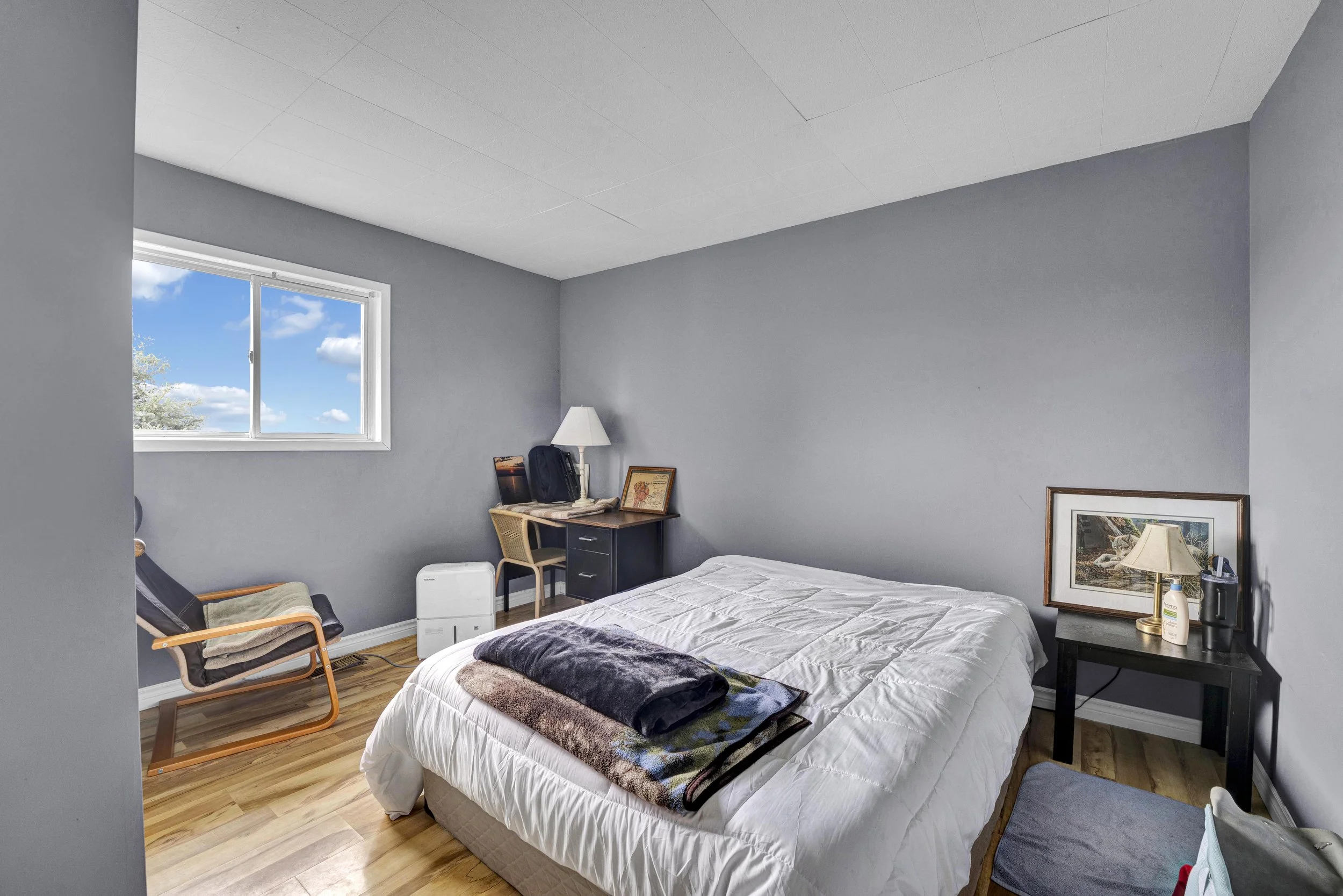 A bedroom with light gray walls, a white ceiling, and a wooden floor. There is a window with a view of blue sky and clouds. A bed with white bedding and a dark blanket is in the foreground. To the right of the bed is a black bedside table with a lamp