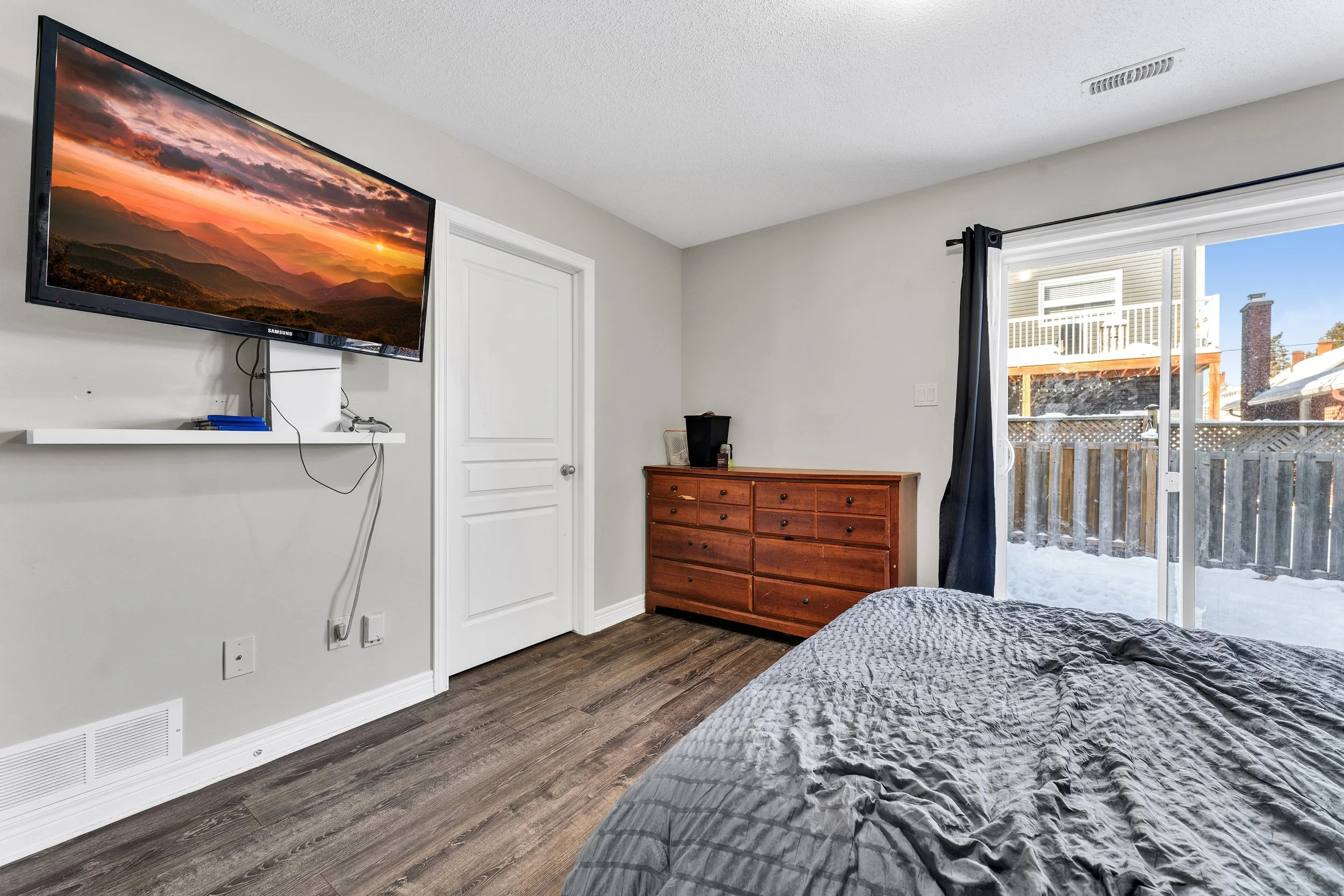 A bedroom with a mounted flat-screen TV on the wall, a wooden dresser, a partially visible bed with gray bedding, and a sliding glass door leading to a snowy outdoor area with a wooden fence and neighboring houses.
