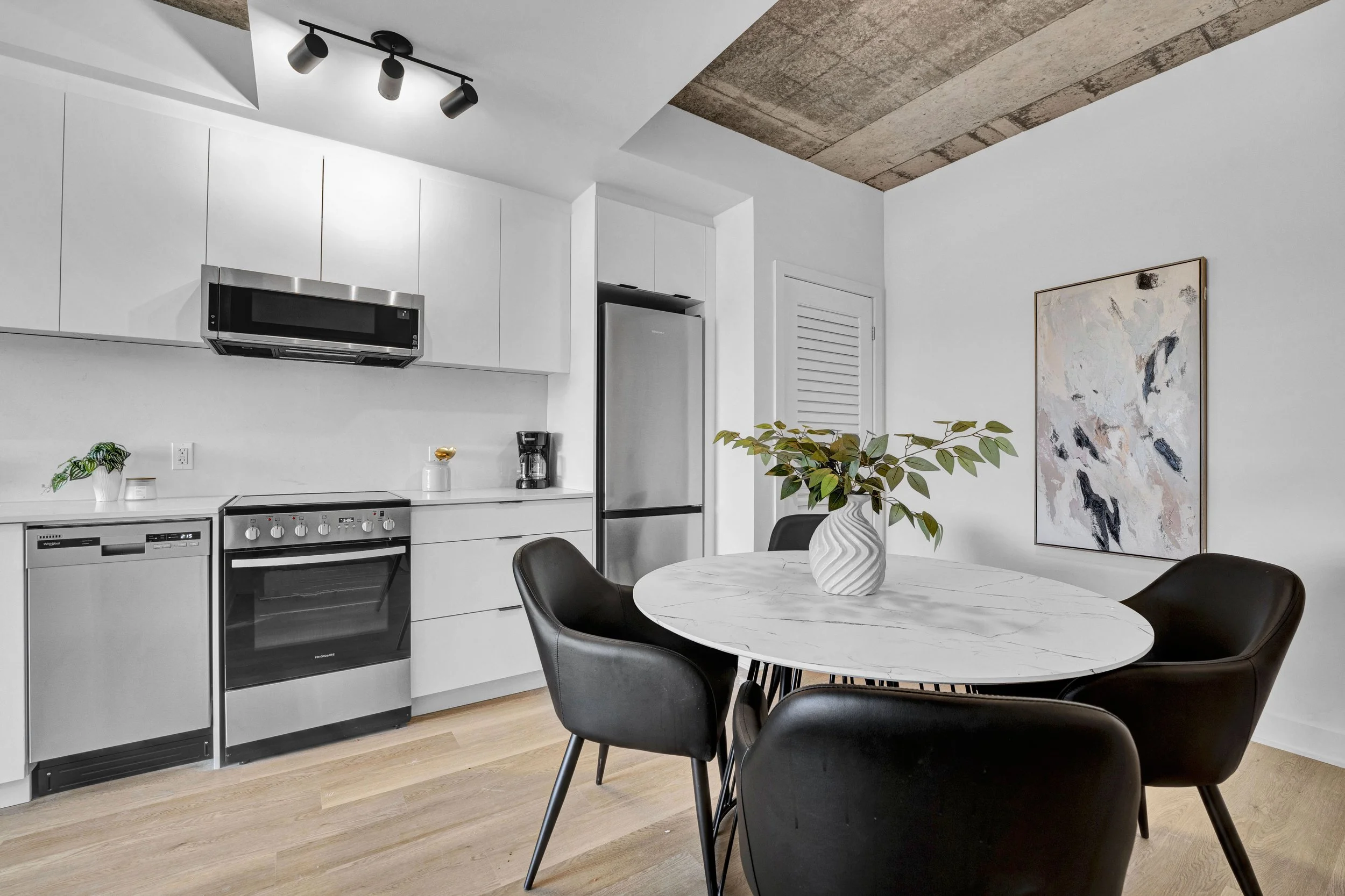 Modern kitchen and dining area with white cabinetry, stainless steel appliances, a round marble table, and four black chairs. There is a decorative vase with green foliage on the table, a plant on the counter, a framed abstract artwork on the wall, a