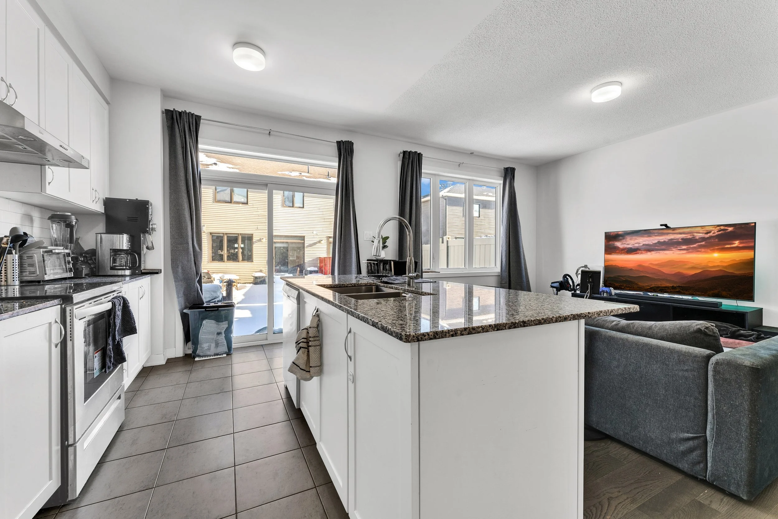 Open-plan kitchen and living room with white cabinets, granite countertops, sliding glass door, and a gray sofa facing a flat-screen TV