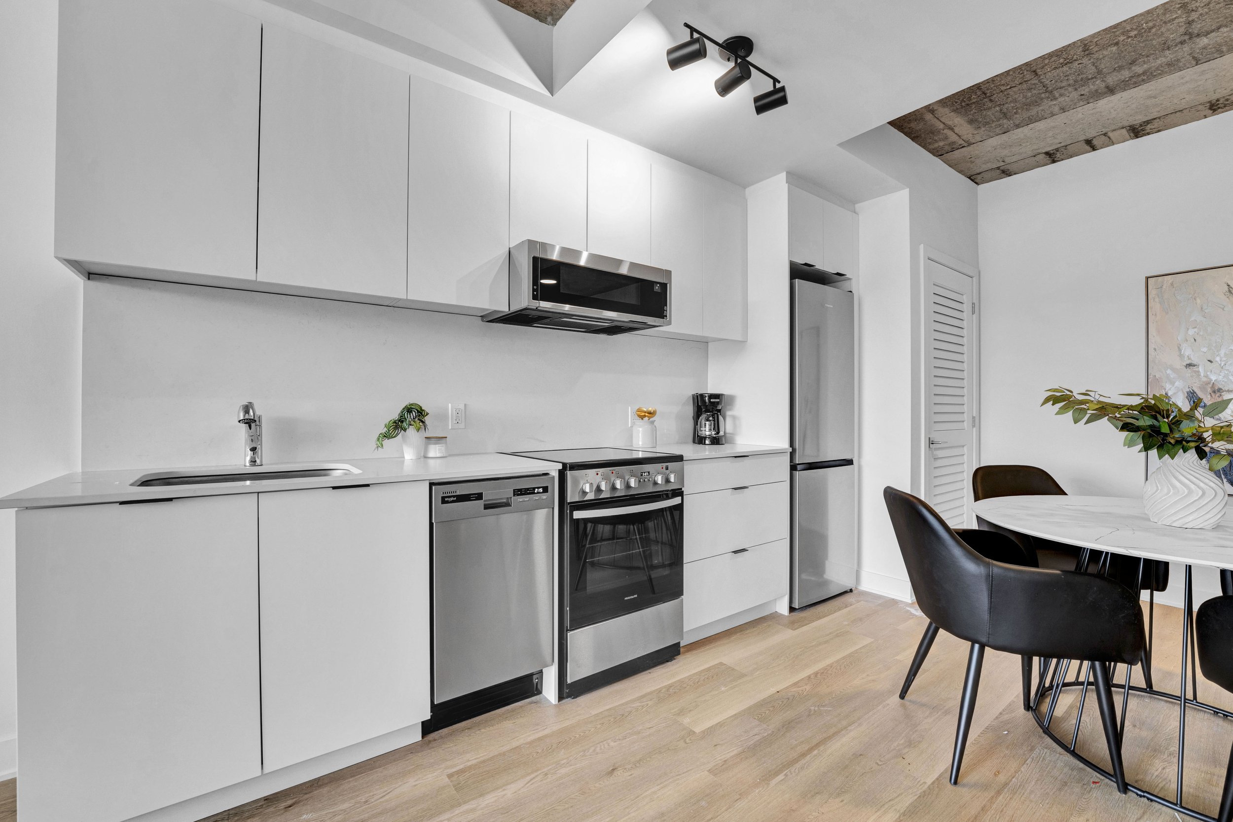 Modern kitchen with white cabinets, stainless steel appliances, black dining chairs, and a round dining table with a plant in a white vase on top.