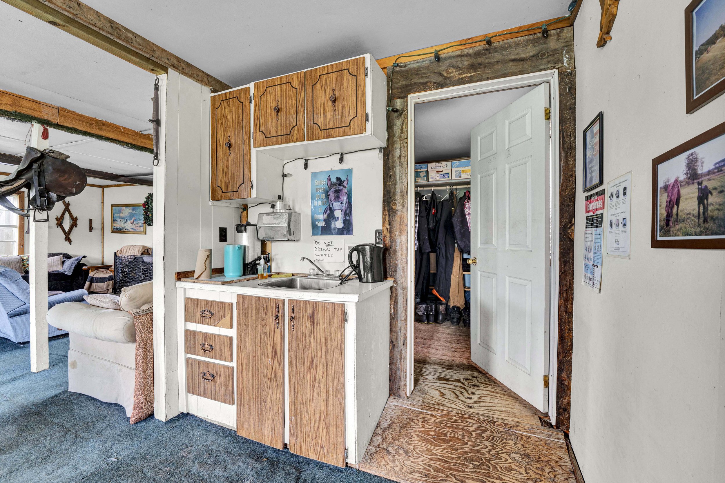 Interior view of a rustic home, showing a small kitchen with wooden cabinets, a sink, and a sign above the sink that reads 'DO NOT DRINK TAP WATER'. In the background, an open doorway leads to a closet with hanging clothes. The walls are decorated wi
