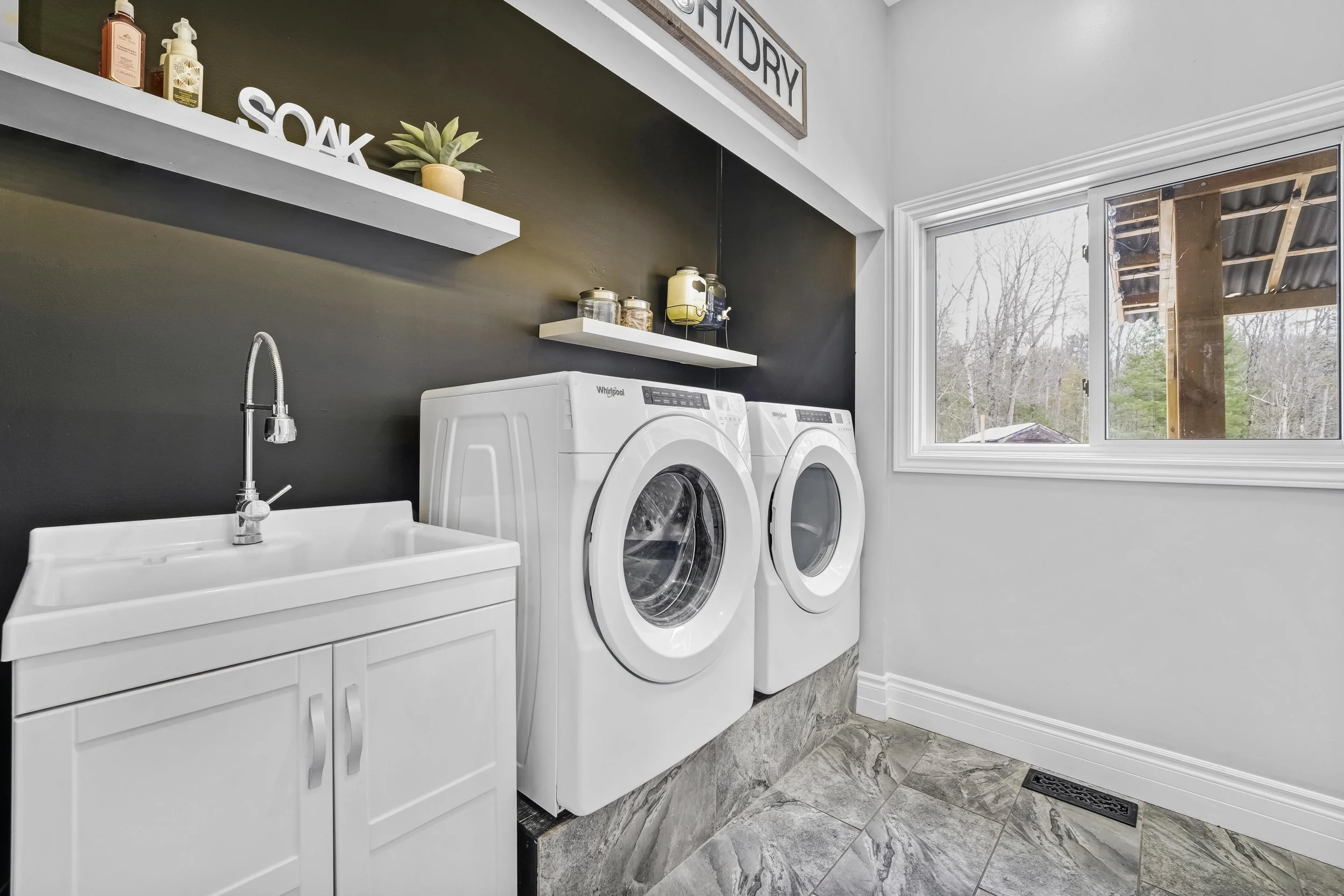 Real Estate Photography in Ottawa Laundry room with washing machine and dryer, white utility sink, black accent wall, white shelves with toiletries and decor, window with view of outdoor construction site.