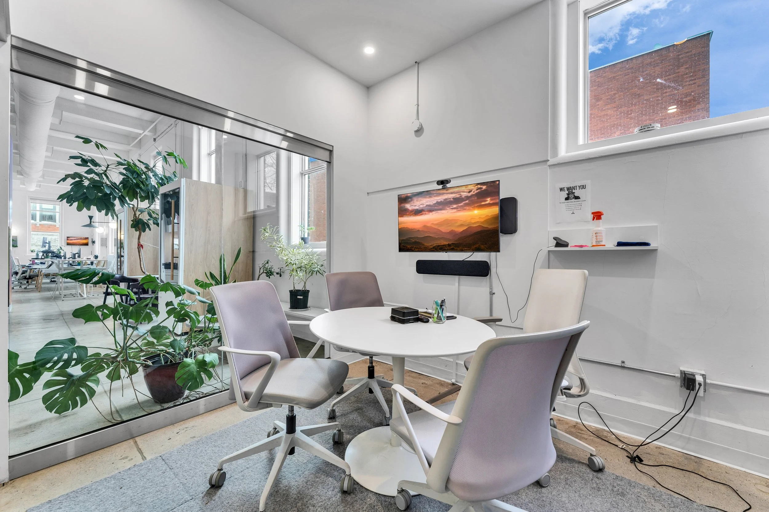 A modern conference room with a round white table surrounded by four white office chairs on casters. The room has white walls, a window, and a large glass partition revealing an adjacent space with more plants and office furniture. On the wall, a tel