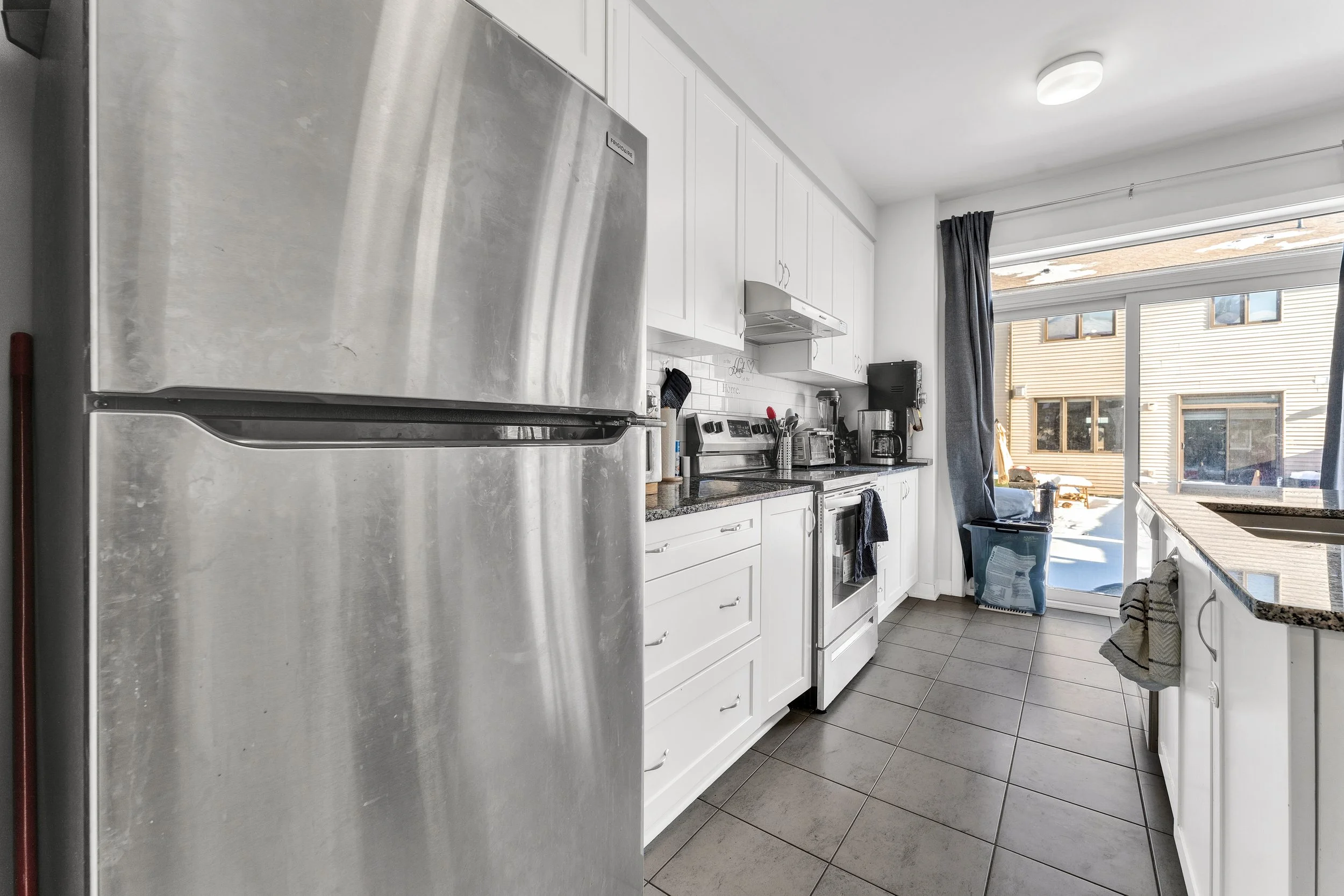 A modern kitchen with stainless steel refrigerator, white cabinets, black countertops, and large window with gray curtains. NV3D Real Estate Photography Ottawa 