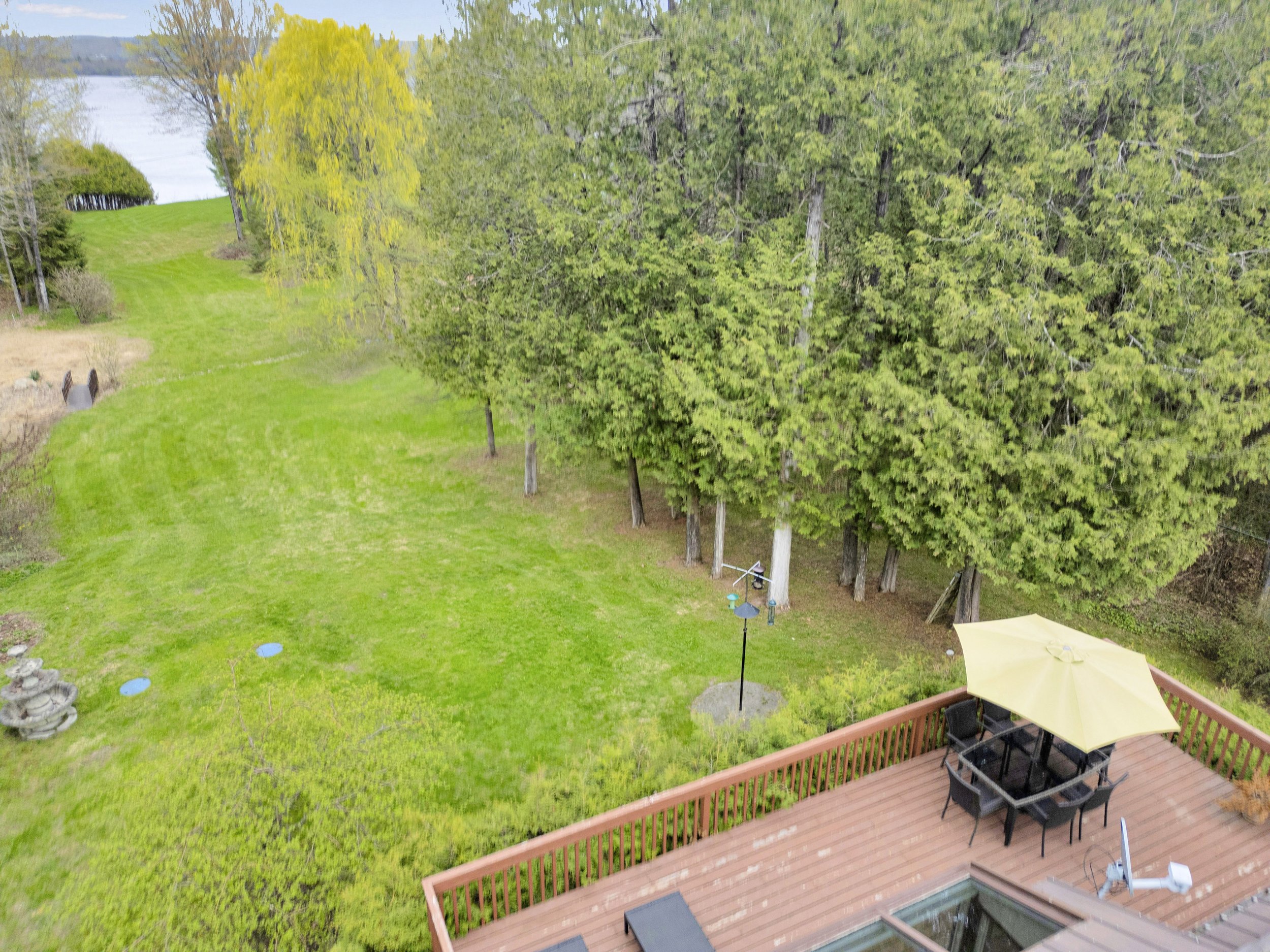 A backyard view featuring a wooden deck with a table, chairs, and a yellow umbrella. The yard has lush green grass, a few decorative stones, and is bordered by tall trees with bright green foliage. The background shows more trees and a view of a rive