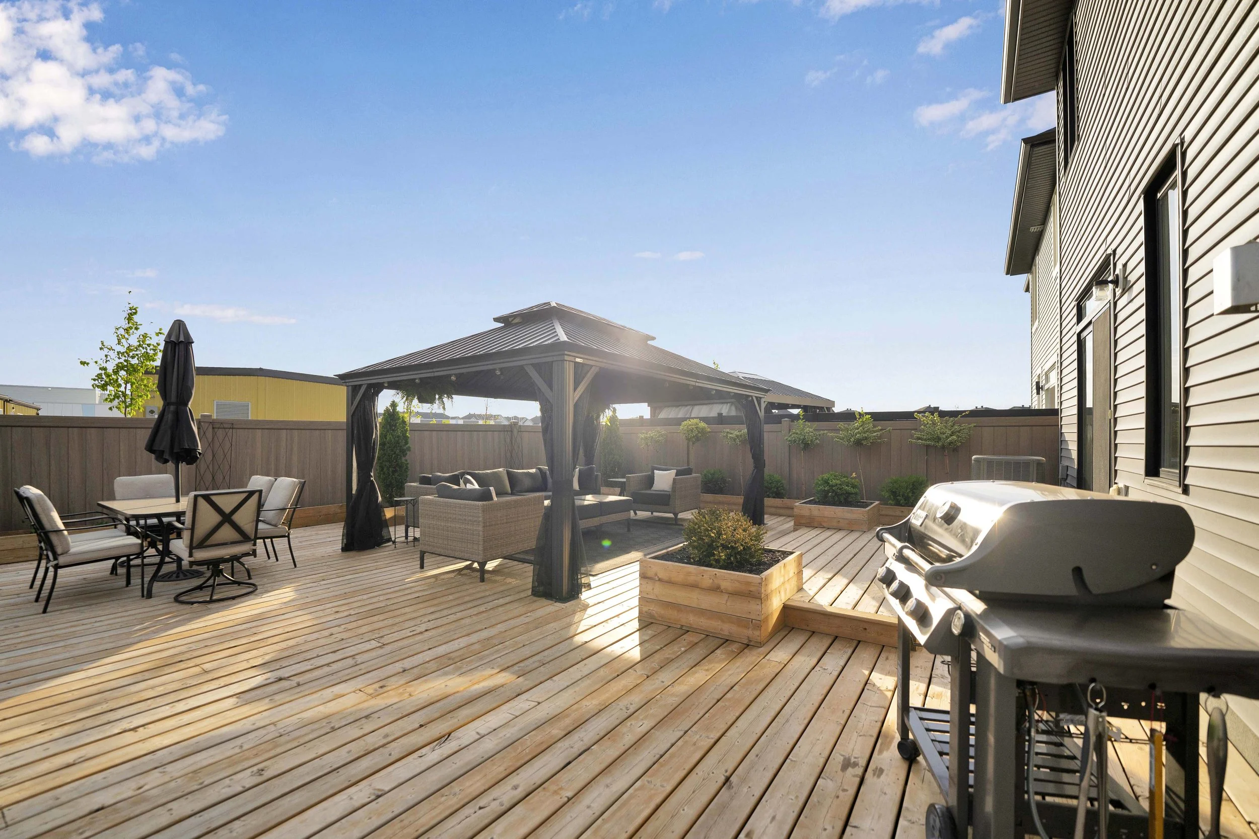 A backyard deck with outdoor furniture, including a dining table with chairs, a barbecue grill, a gazebo with seating underneath, and decorative potted plants, surrounded by a wooden fence and a clear blue sky.