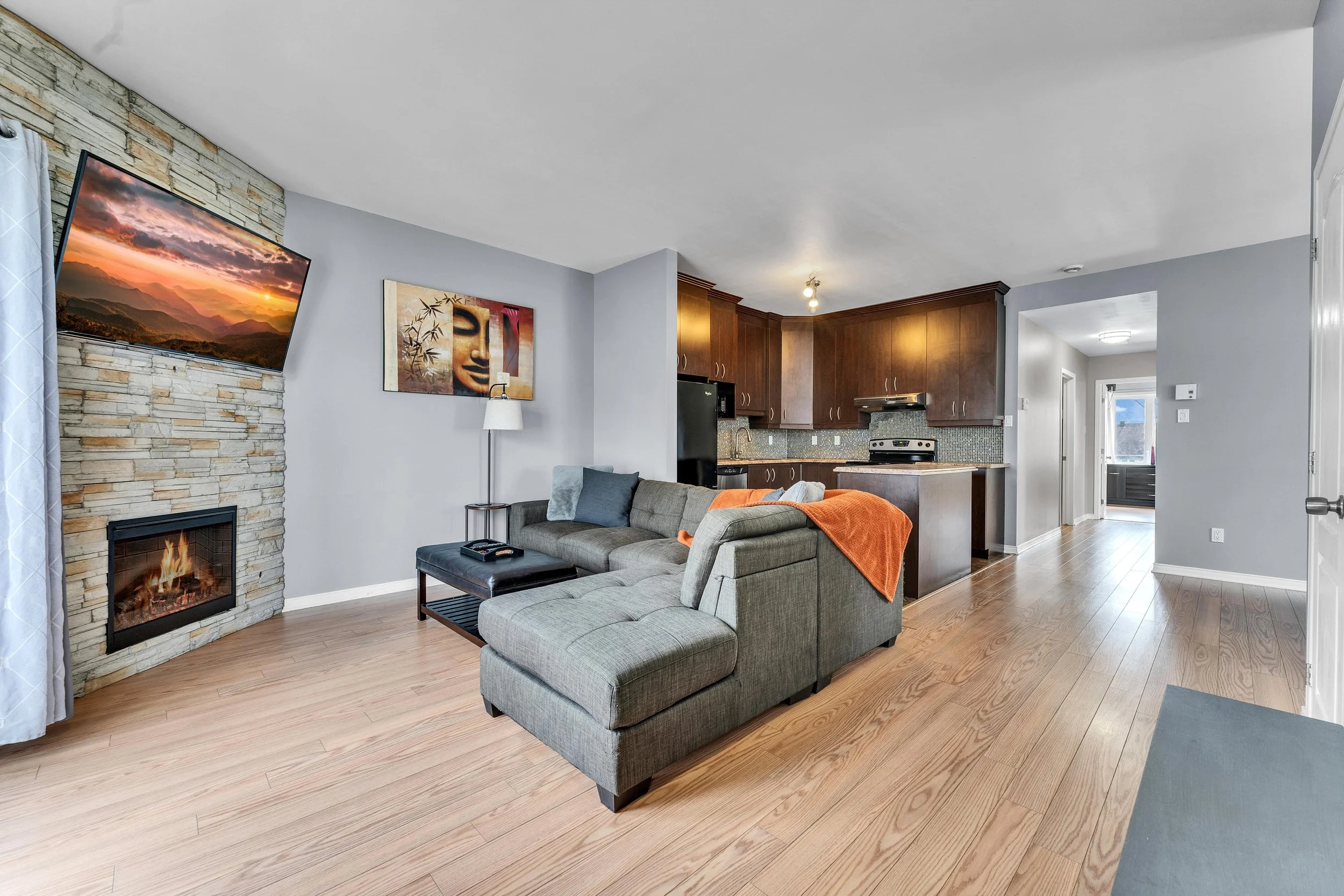 Open-concept living room and kitchen with a sectional sofa, wall-mounted TV, stone fireplace, artwork, wooden cabinets, and hardwood floors.