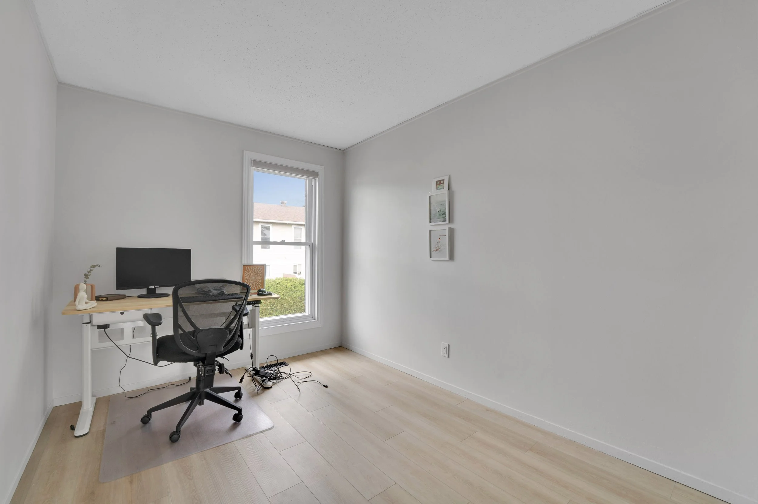 NVISION3D Real Estate Photography Ottawa  A minimalist home office with a small desk, a black ergonomic chair, a computer monitor, and a window with a view of neighboring houses and blue sky. The walls are white, and the flooring is light wood.