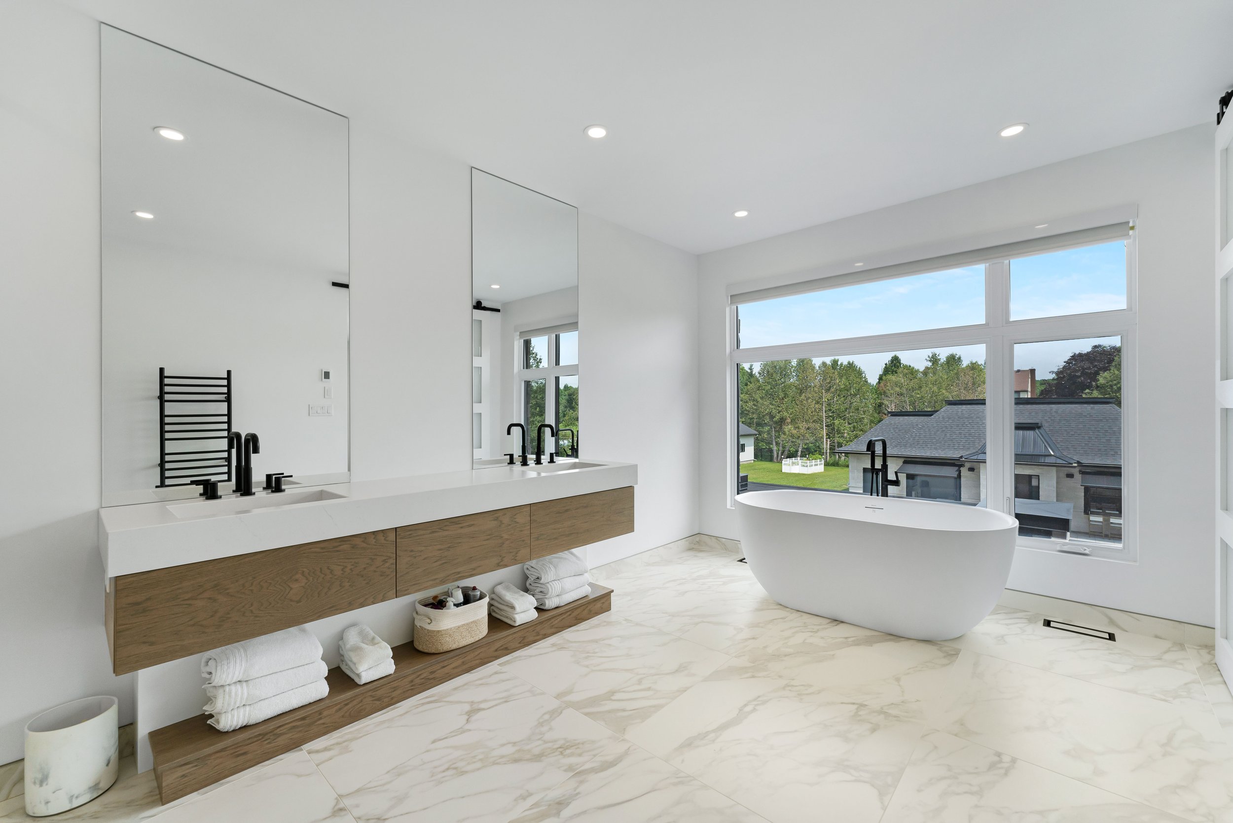 Modern bathroom with freestanding bathtub, large window, dual sink vanity with mirrors, and marble tile floor. Real estate photography ottawa