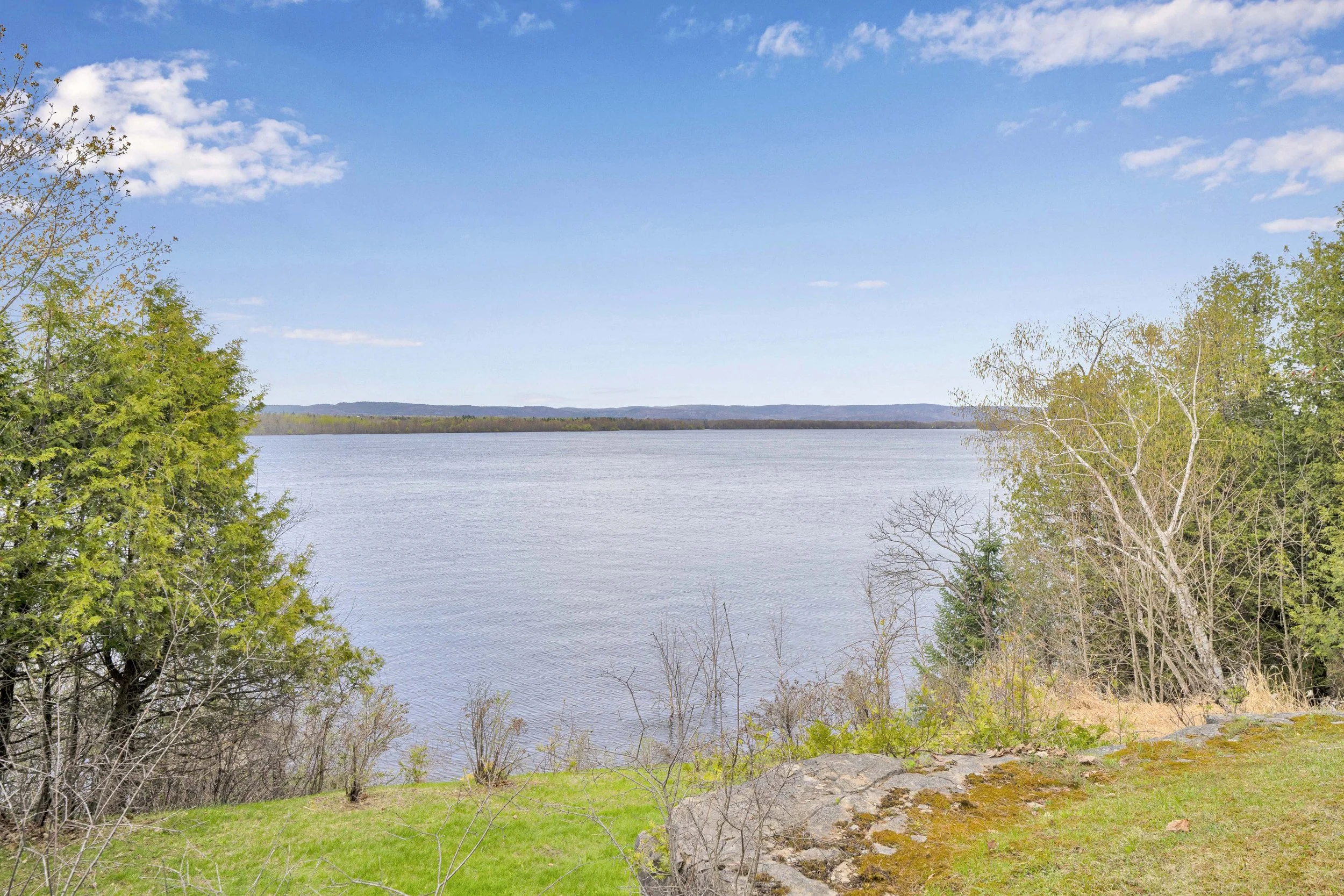 NVISION3D.ca Real Estate Media Ottawa  Scenic view of a river or lake with trees and hills in the background under a partly cloudy blue sky.