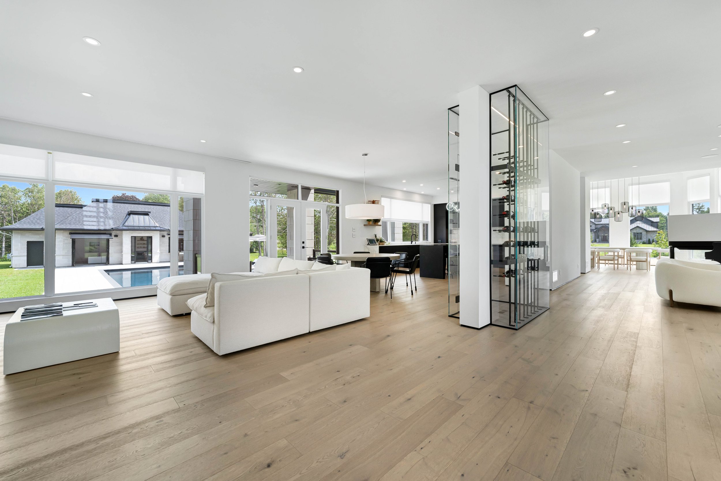Modern open-plan living area with white sofas, wood flooring, dining area with black chairs, glass wine storage, large windows showing outdoor pool and small building.