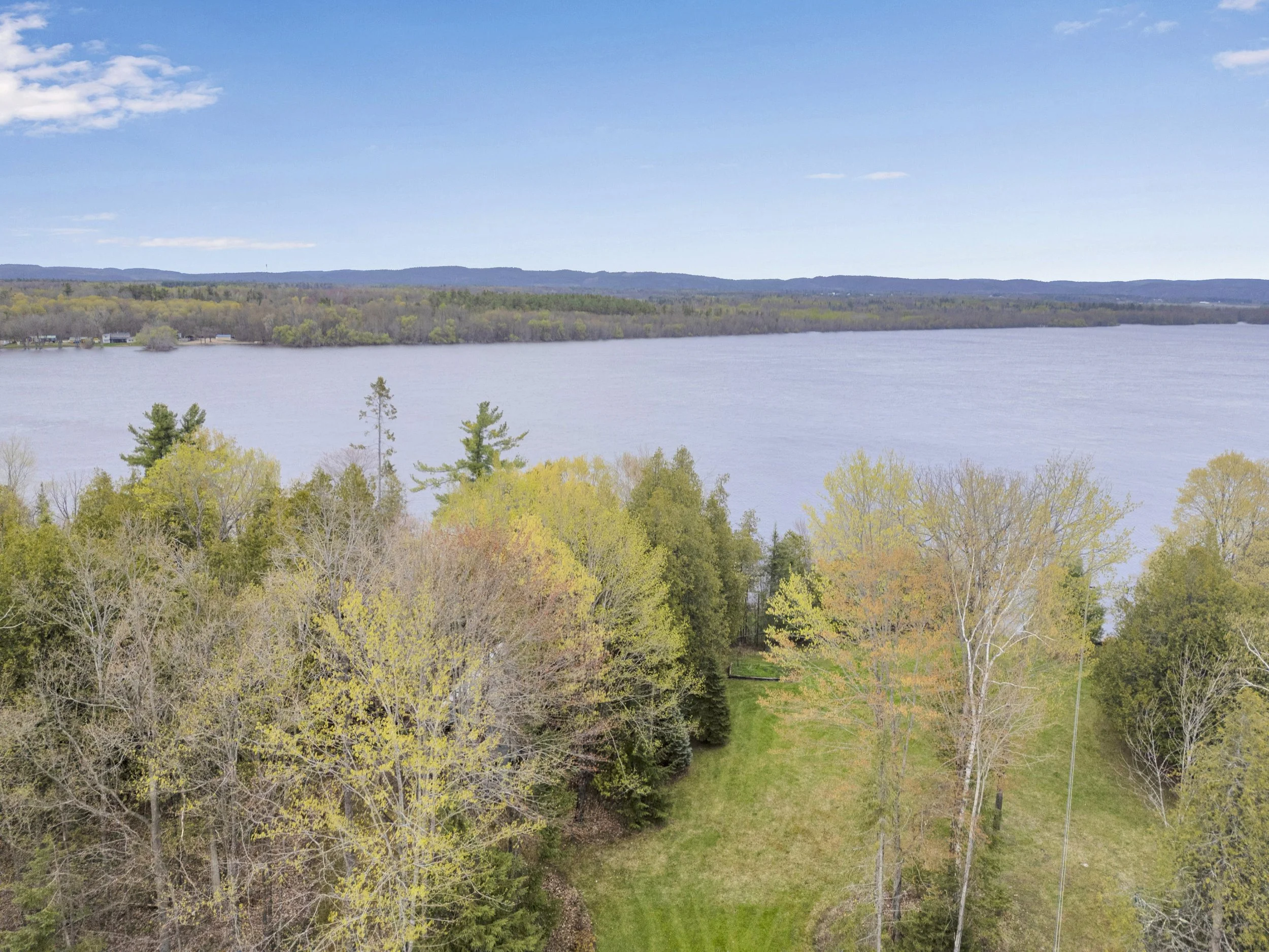 Ottawa Real Estate Media Services  A landscape with a river in the foreground, surrounded by trees with green and early spring foliage, and hills in the background under a partly cloudy sky.